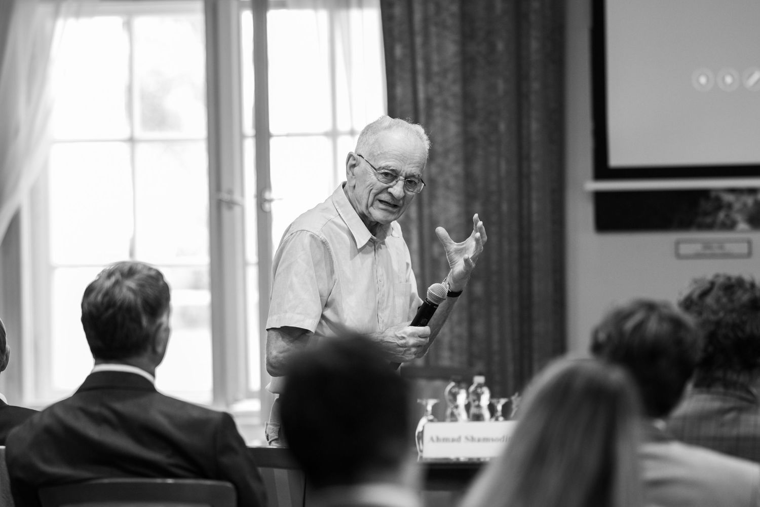 Black and white photograph of a person gesturing while speaking to an audience in what appears to be a meeting room.