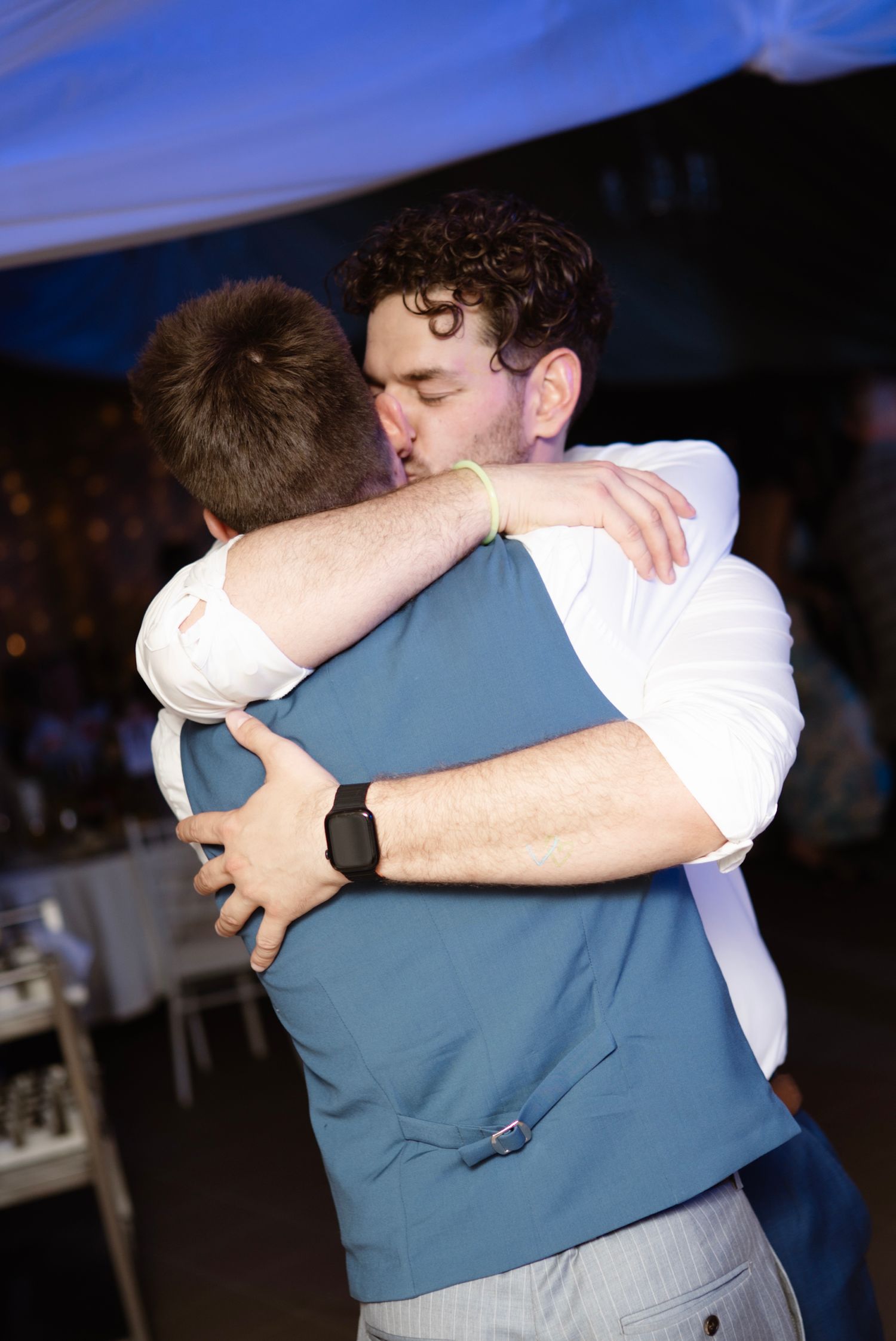 Two men share an emotional hug at a wedding reception under a draped tent.