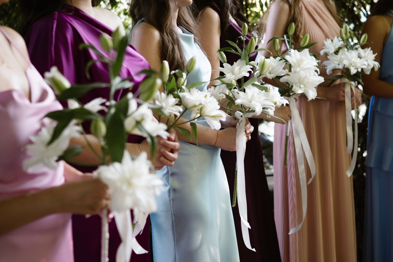 Bridesmaids in colorful dresses holding white lily bouquets lined up at a wedding ceremony.