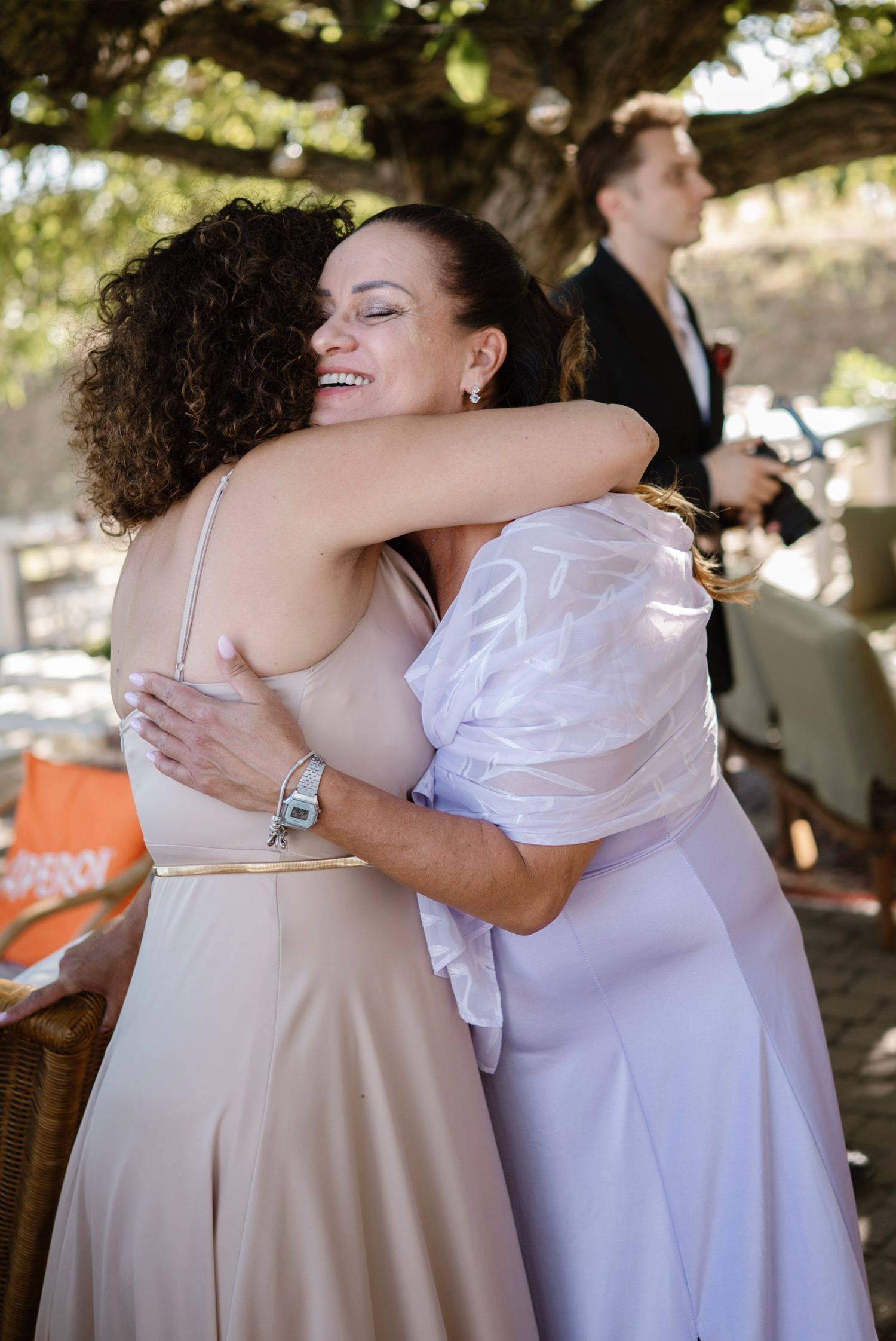 Two women share a warm embrace at an outdoor wedding celebration beneath a shady tree.