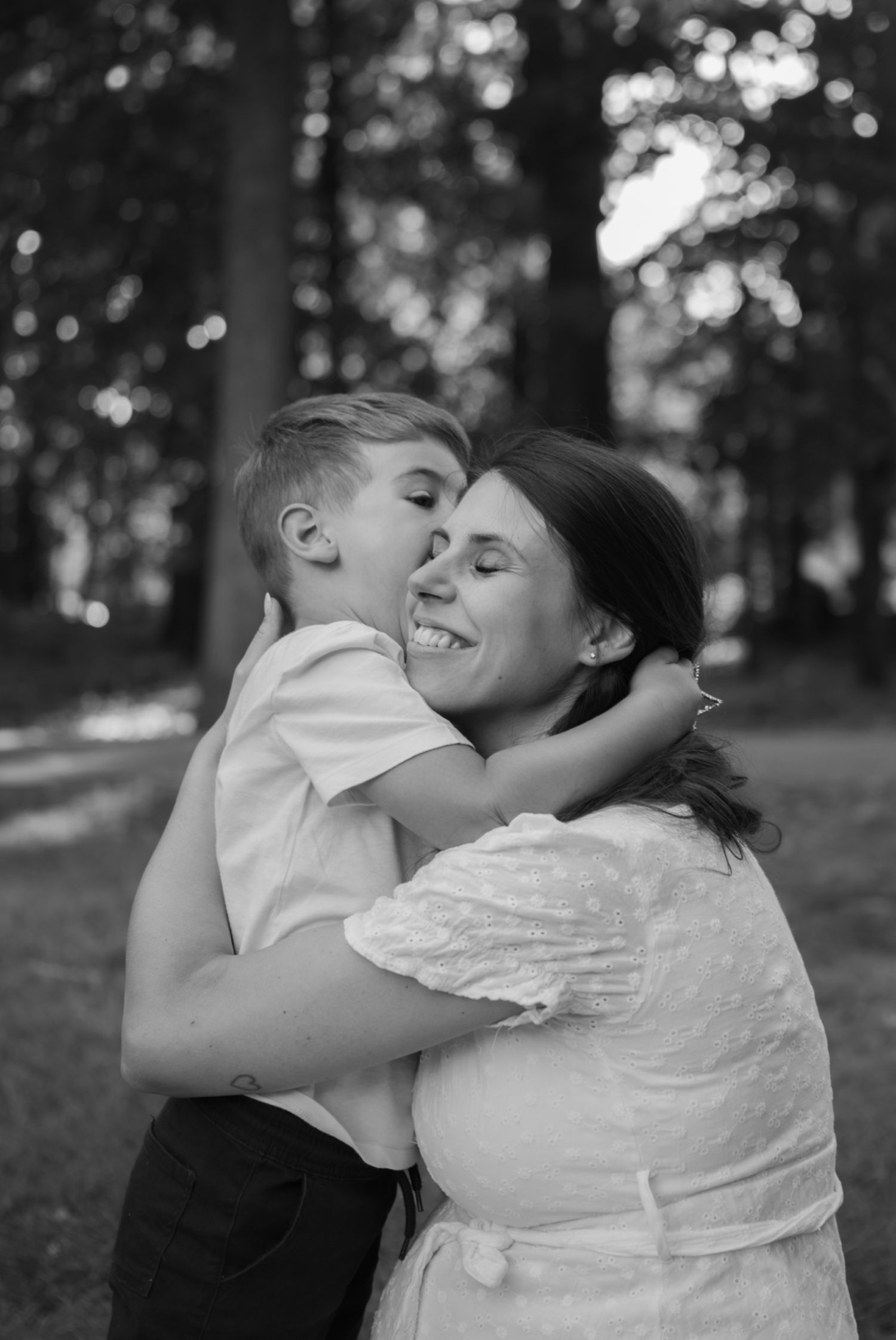 Black and white photo of a young boy hugging and kissing a smiling woman outdoors in a park setting.