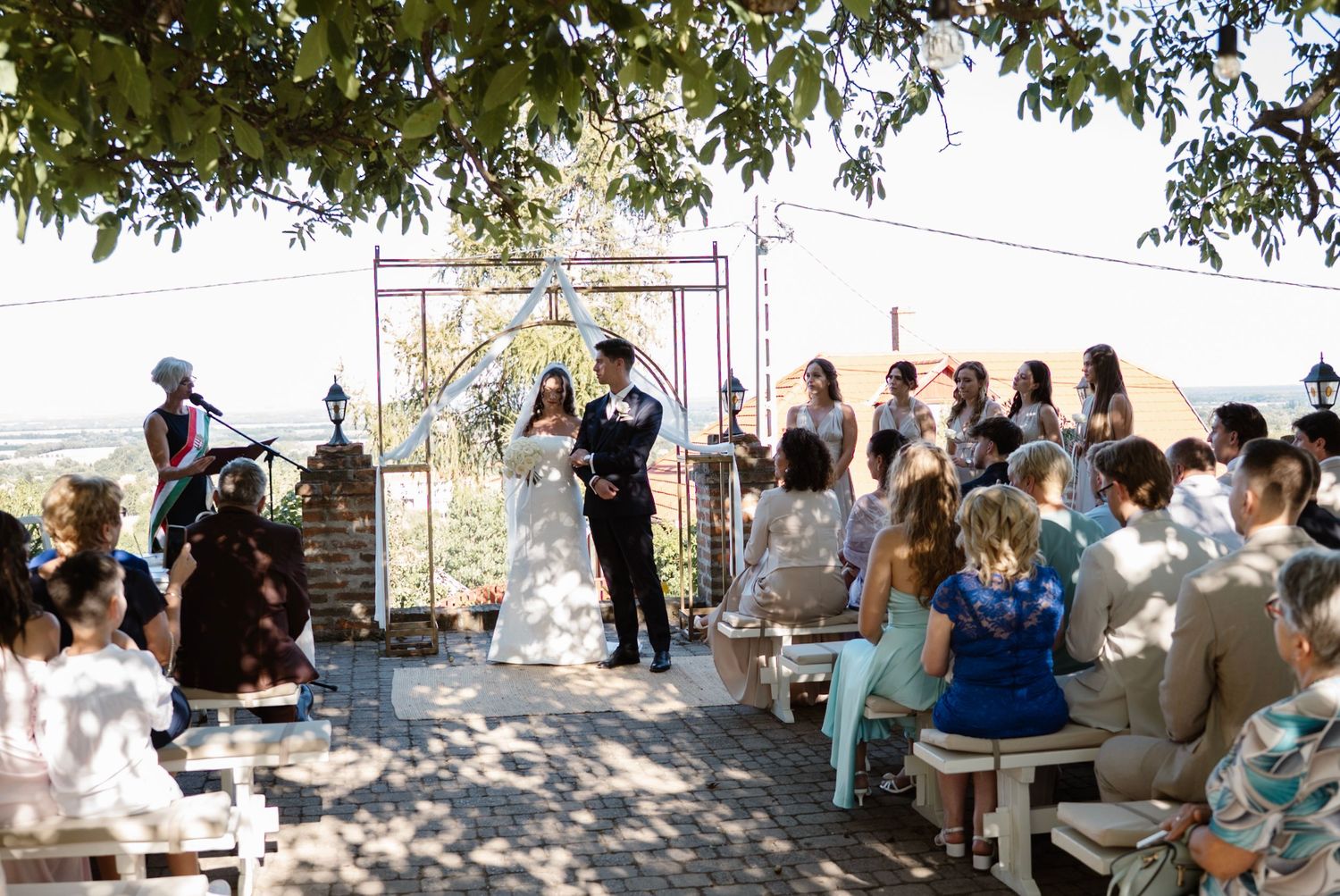 Outdoor wedding ceremony with couple at floral arch, guests seated on white benches under trees.