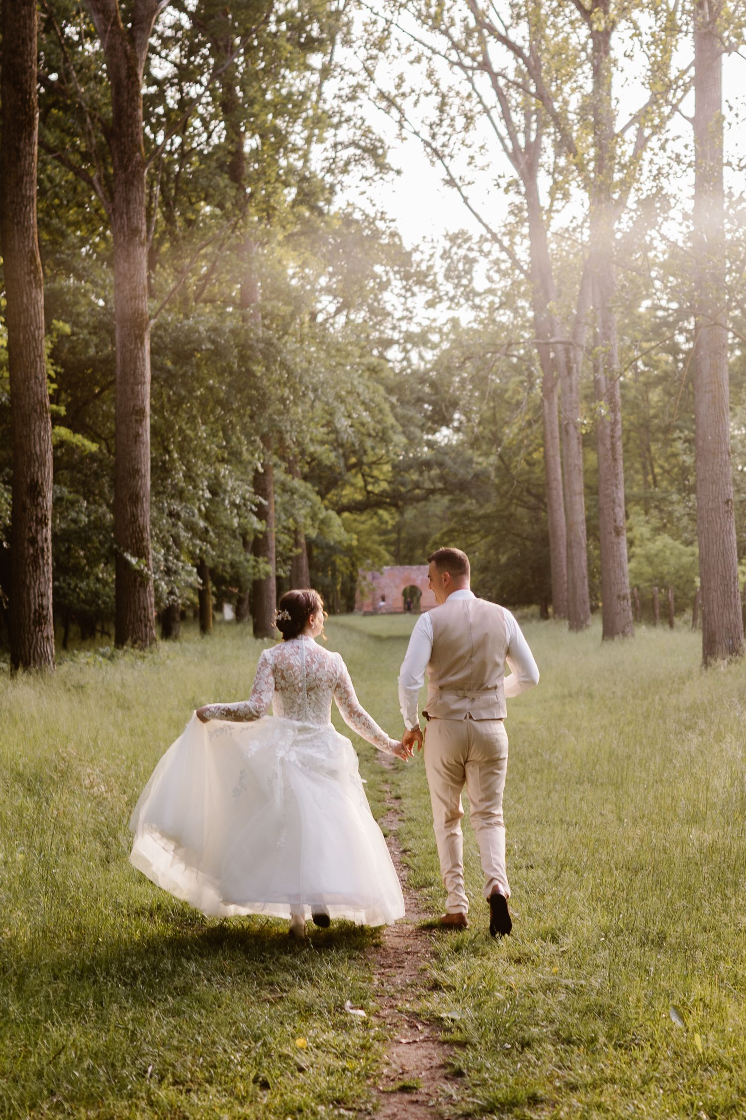 Newlyweds walk hand in hand through a sunlit wooded area in their wedding attire.