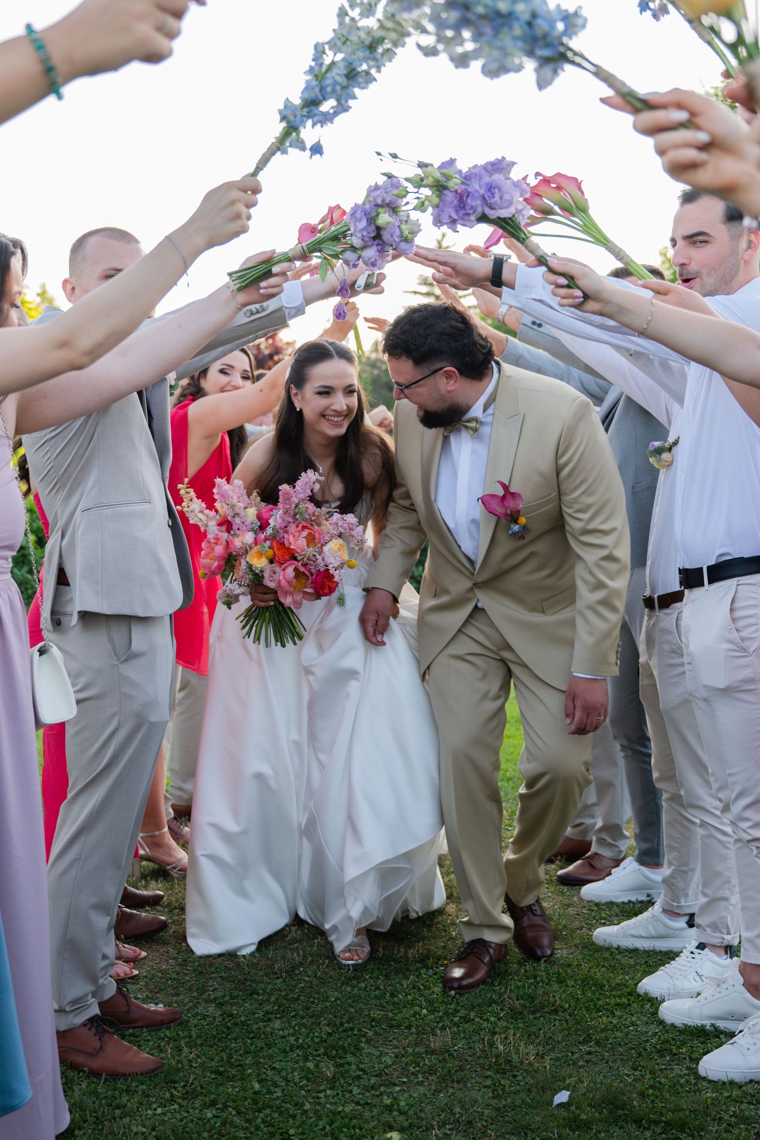 Wedding guests form a tunnel with raised arms for the newlyweds to walk through on a sunny outdoor lawn.