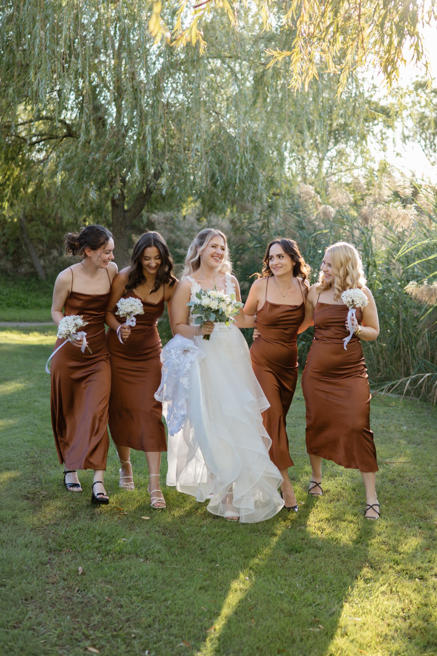 Wedding party in brown dresses and white gown pose together under willow trees at sunset.