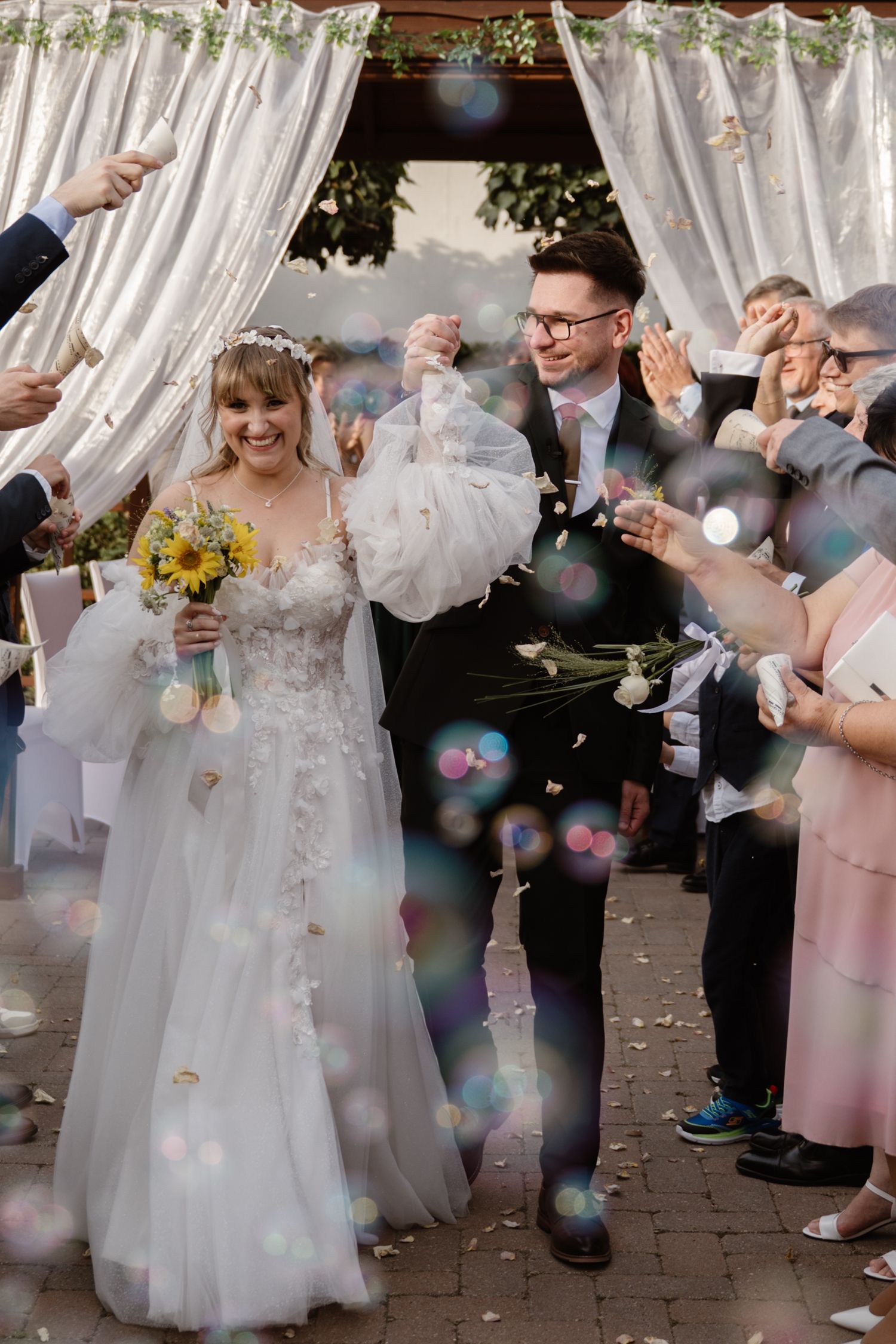 Wedding celebration with bubbles as couple in white dress and guests gather under decorative canopy.