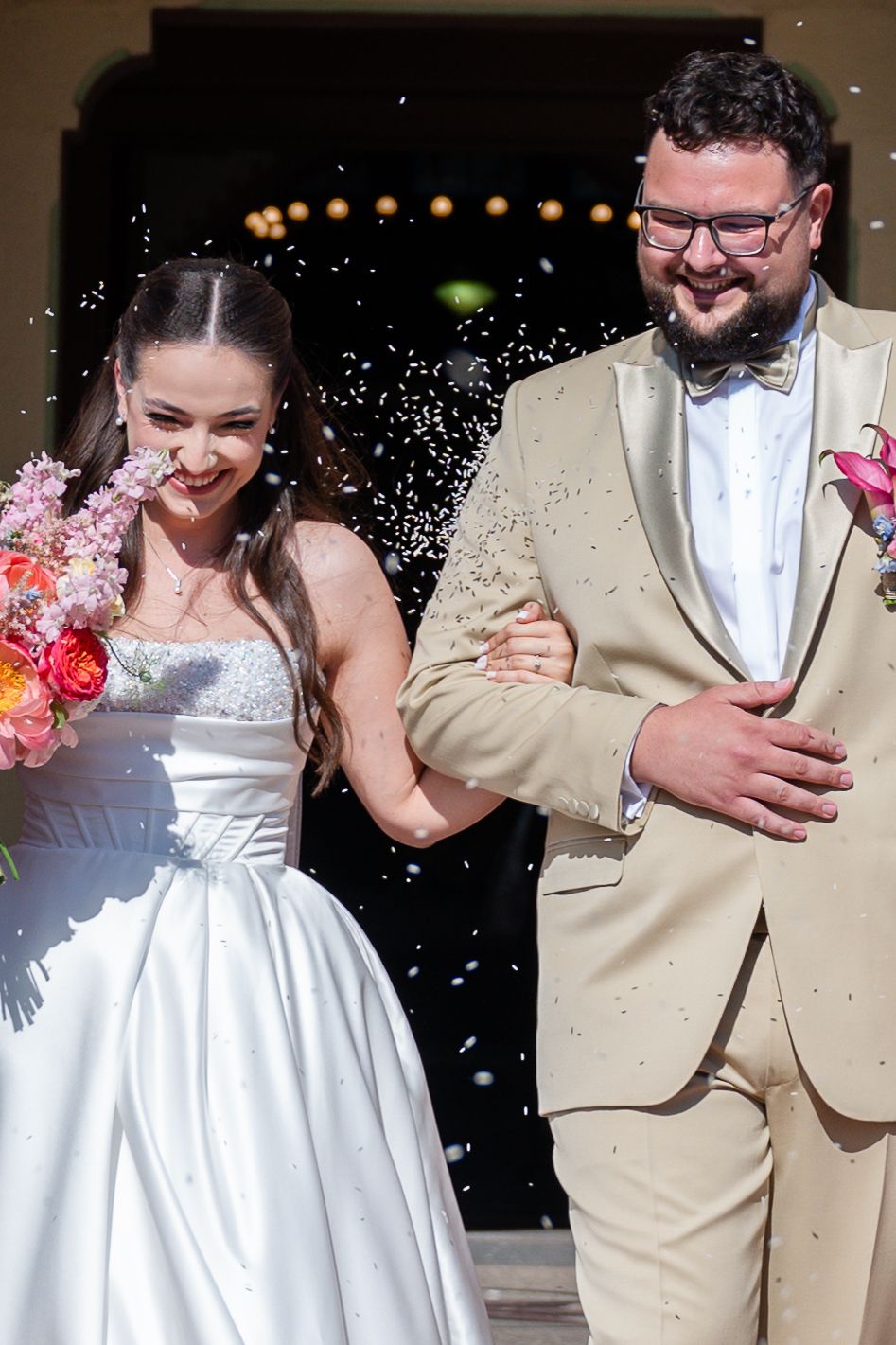 Newlyweds exit their ceremony while being showered with confetti, bride holding colorful bouquet.