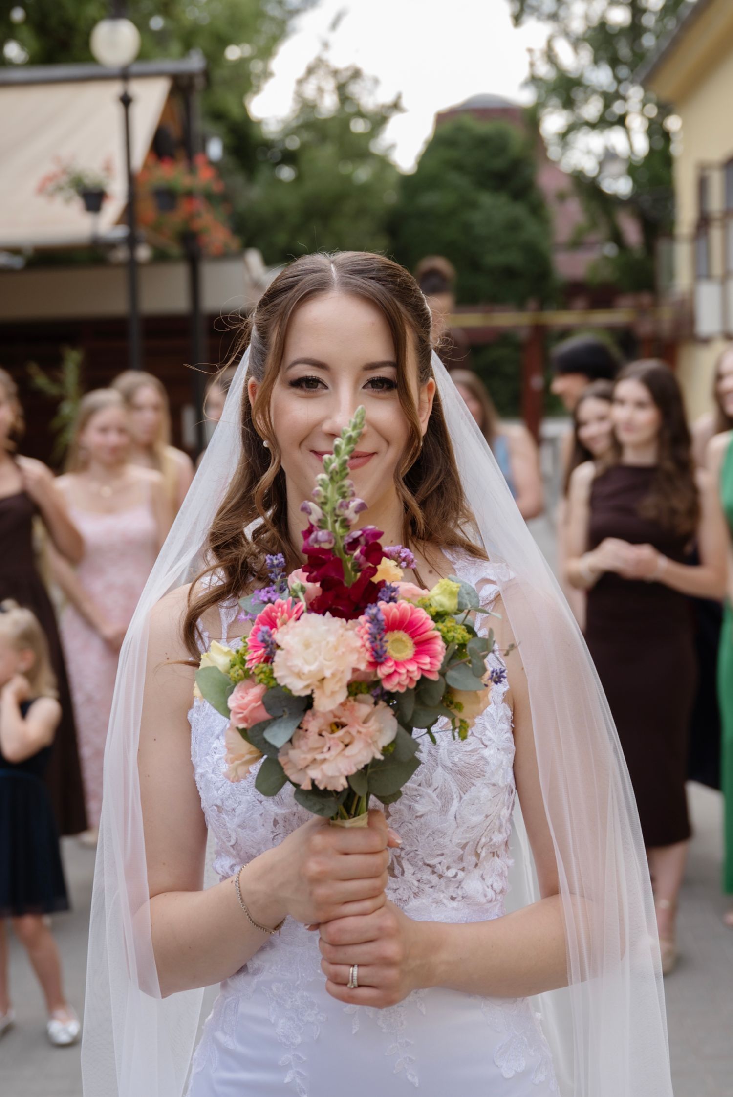 Beautiful bride in white lace gown and veil holds colorful bouquet of pink and red flowers outdoors.