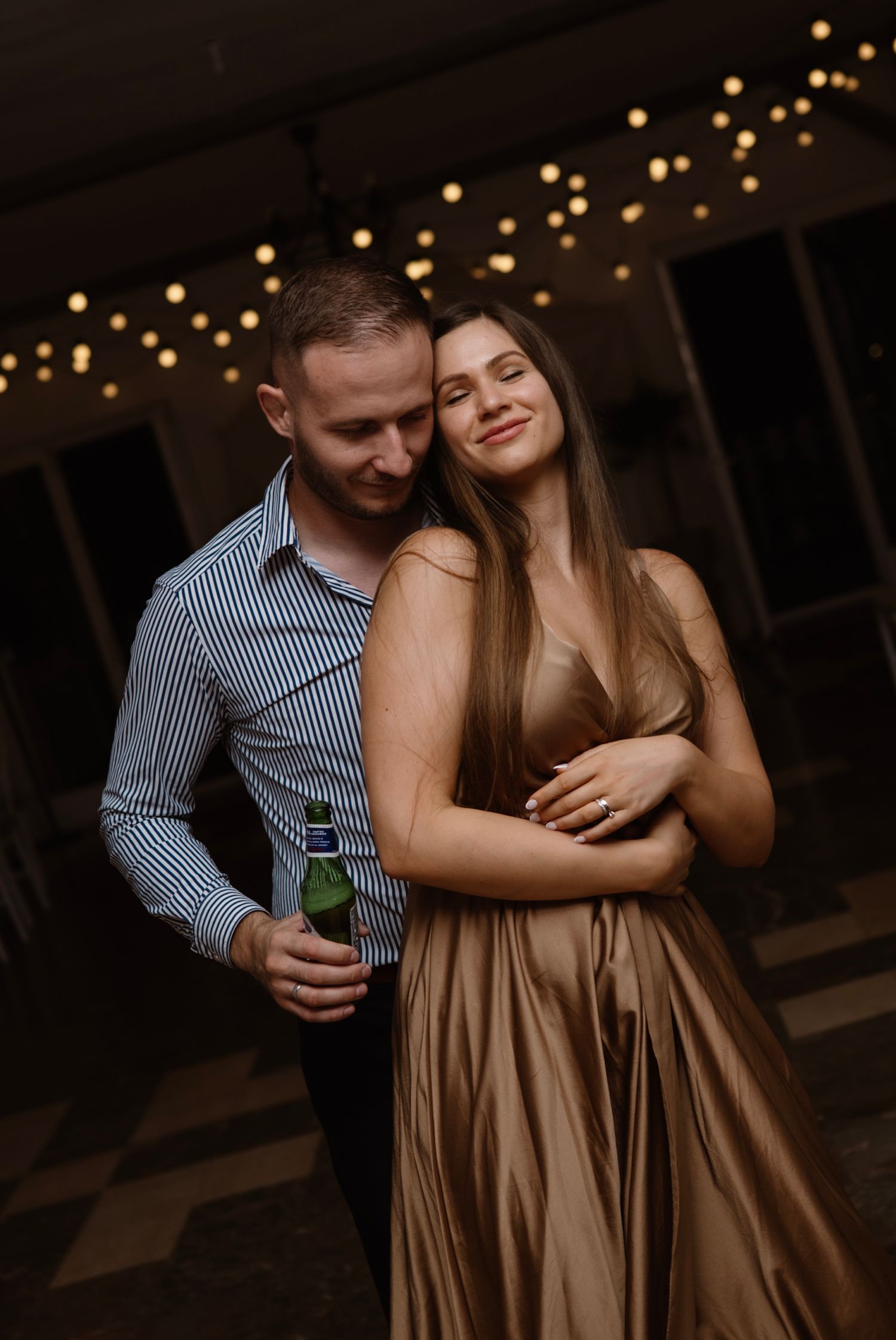 Couple embracing at evening event, woman in gold dress and man in striped shirt holding beer bottle, string lights background.