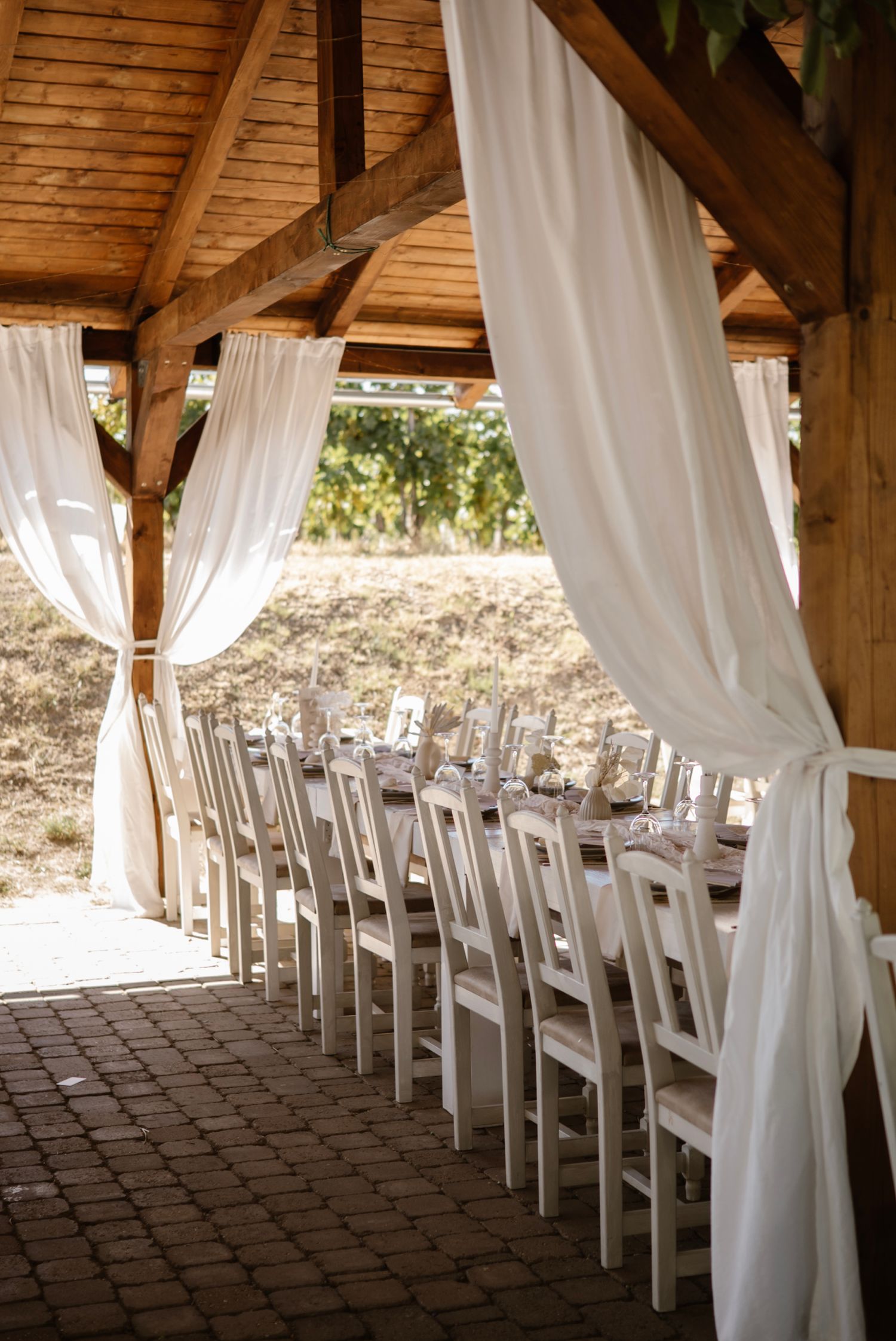 Rustic outdoor wedding reception under wooden pavilion with white draping curtains and long dining table setup.