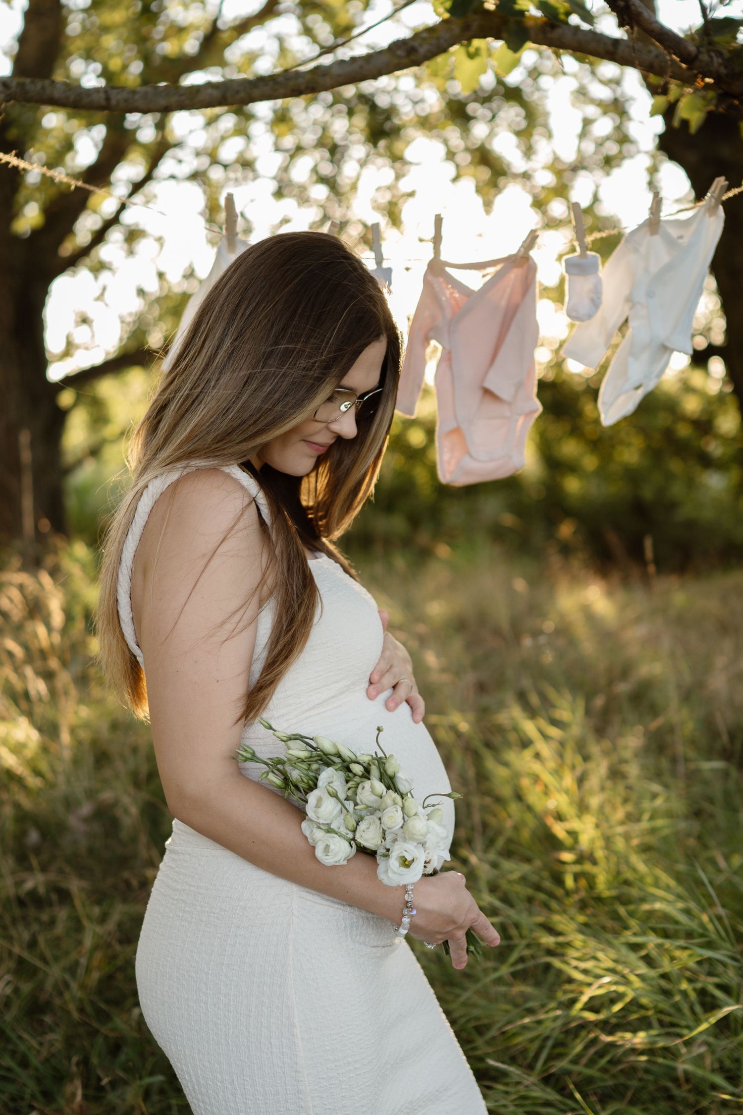 Maternity photoshoot in a field at sunset with white baby clothes hanging on a clothesline.