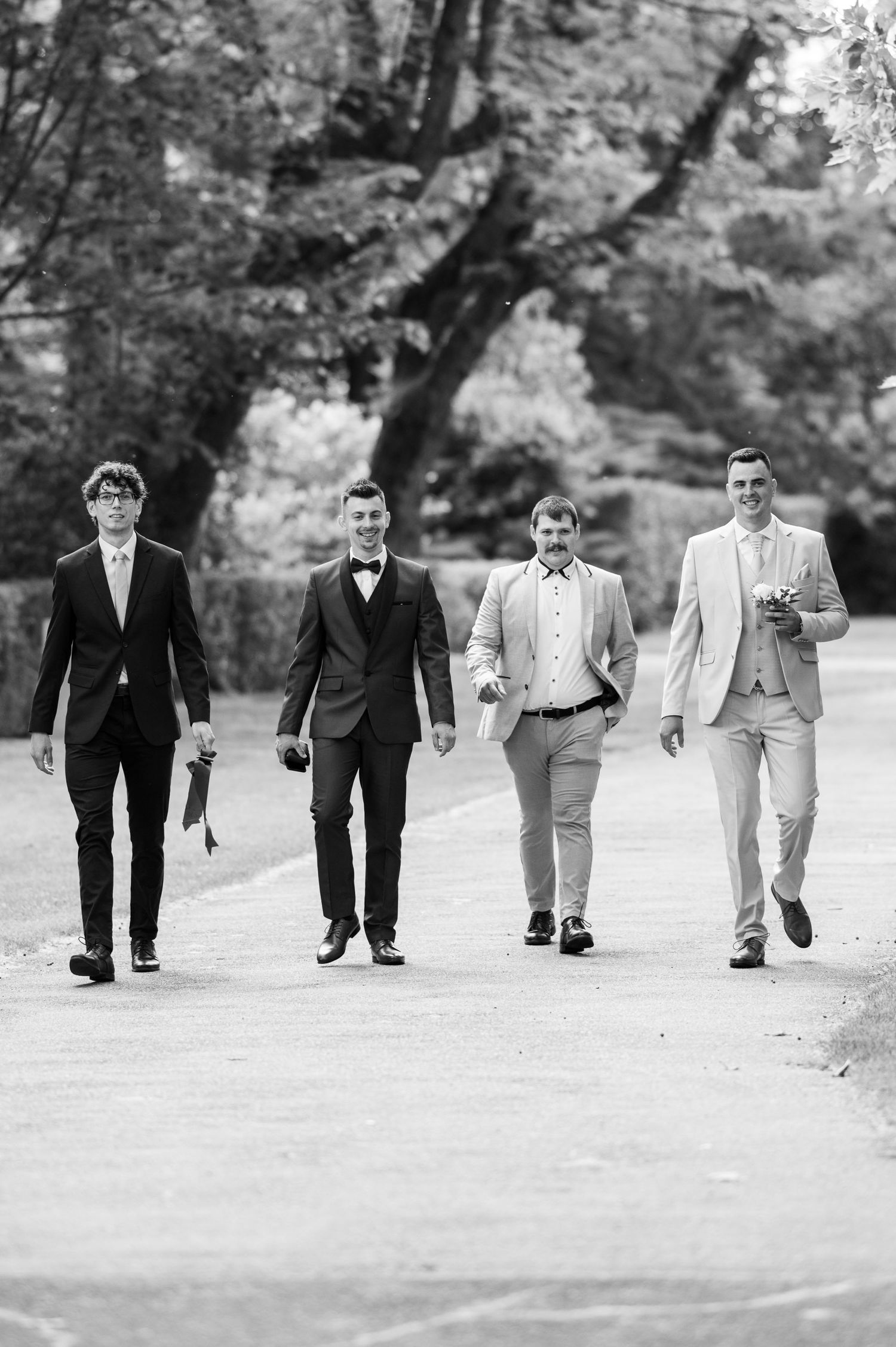 Black and white photo of groomsmen walking in formation on a tree-lined path.