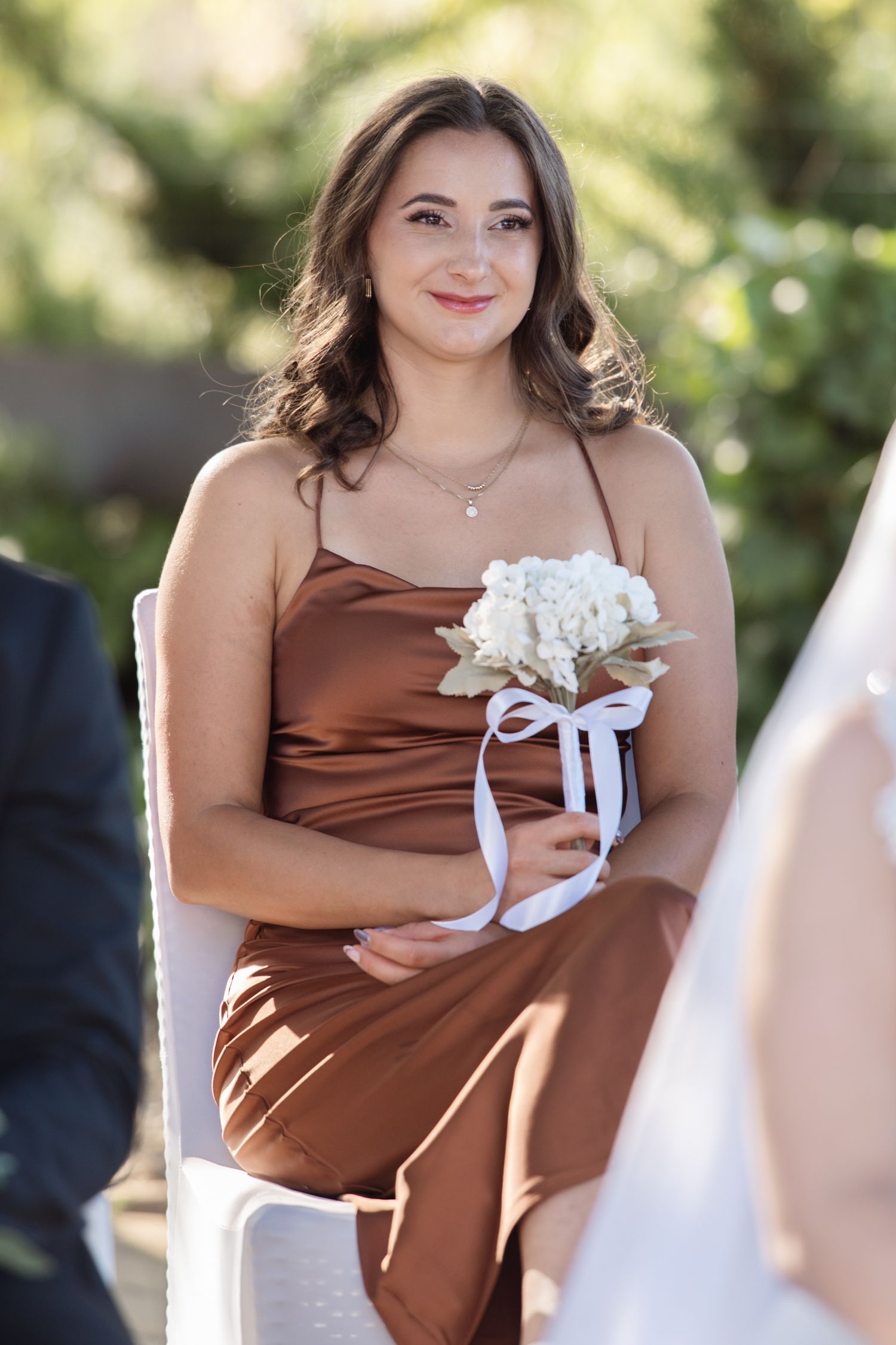 Bridesmaid in a brown satin dress holds a white floral bouquet while sitting in a chair outdoors.