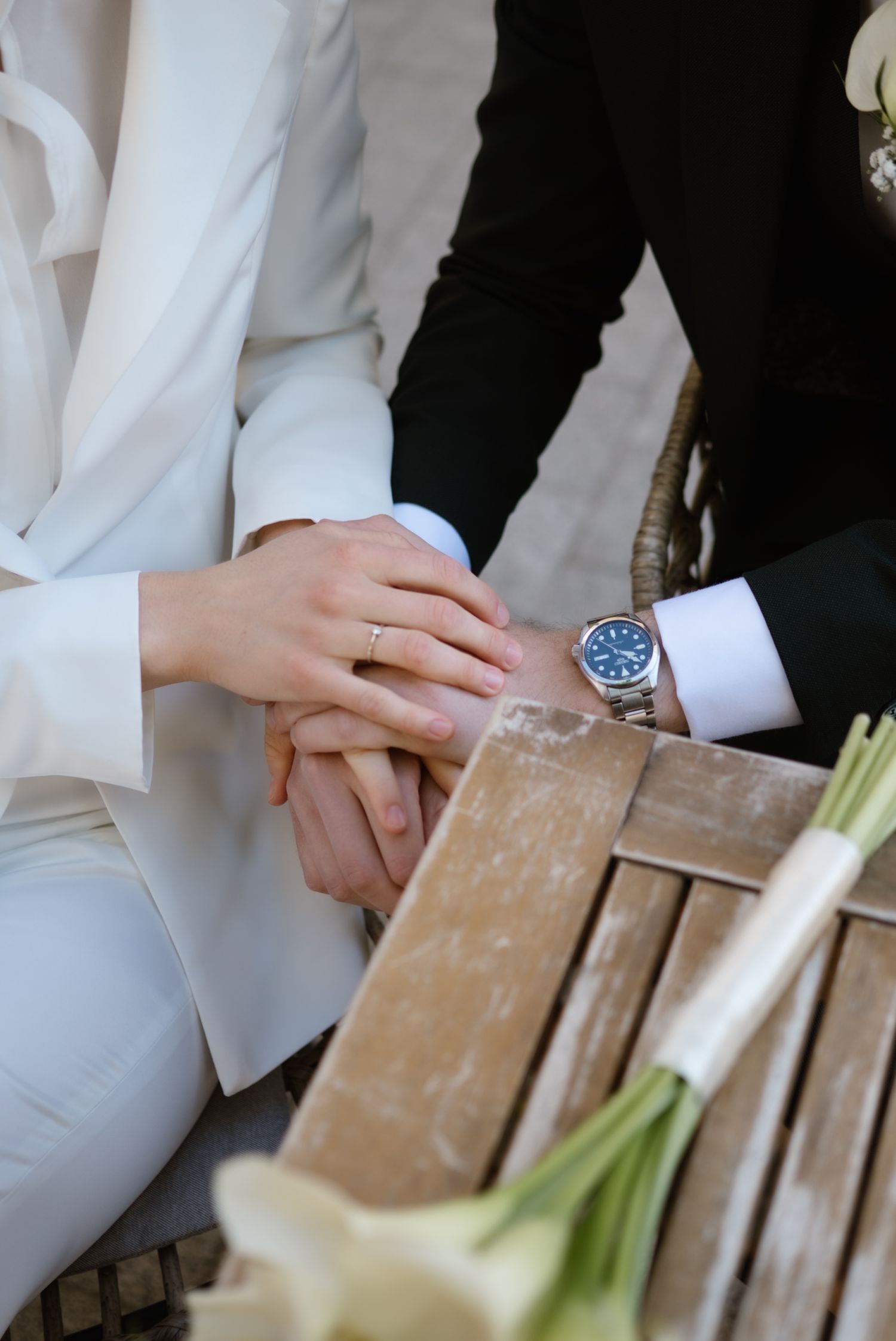 Newlywed couple holding hands on a wooden bench, bride in white suit with gold ring, groom in black suit with blue watch.