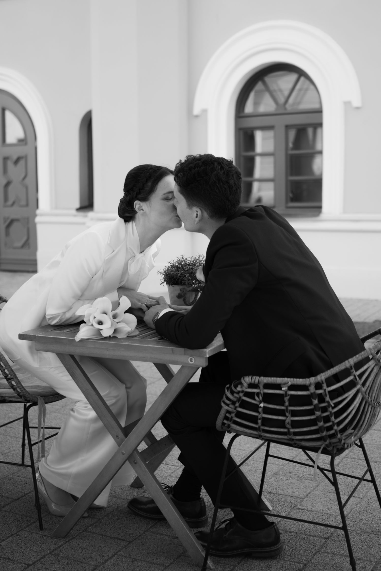 Black and white photo of a couple kissing at an outdoor cafe table, bride in white dress holding flowers.