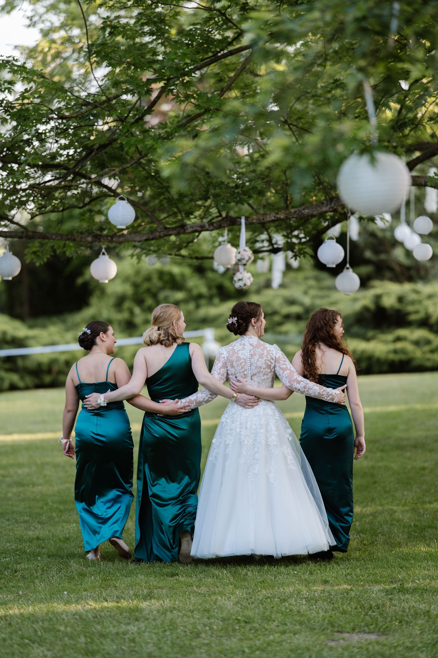 Bridesmaids in teal dresses sit with bride on grass under white paper lanterns.
