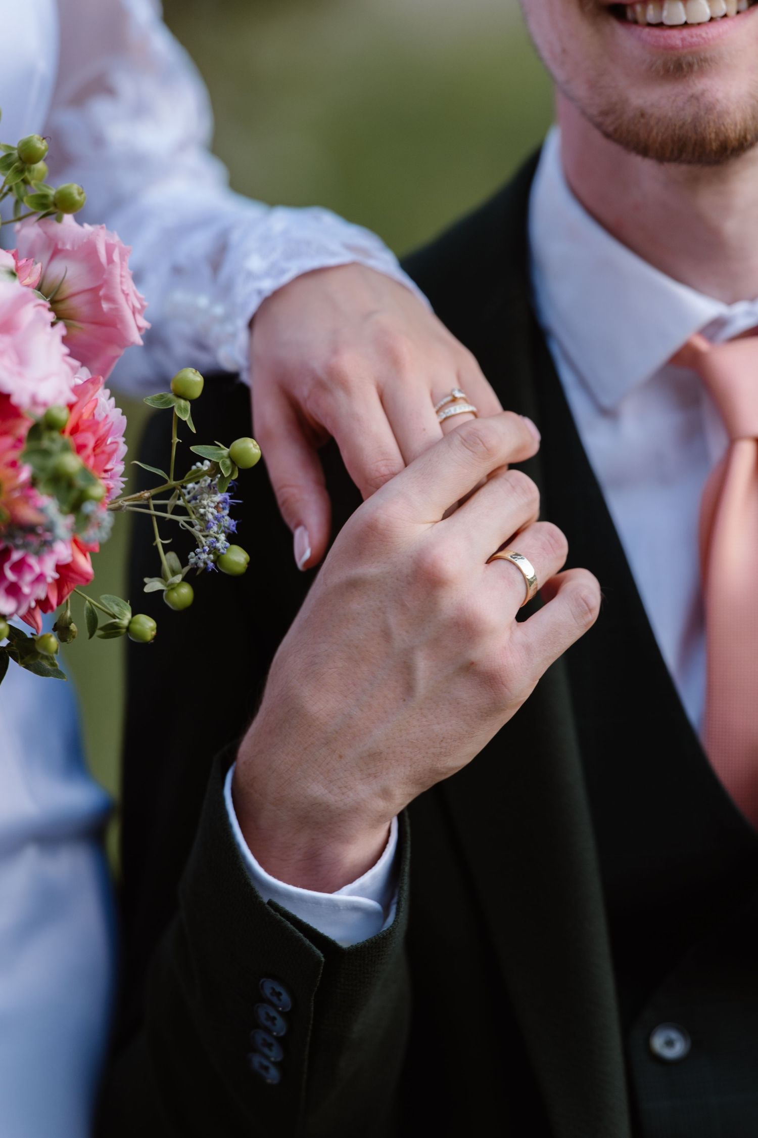 Wedding detail shot showing rings and pink floral bouquet against formal attire.