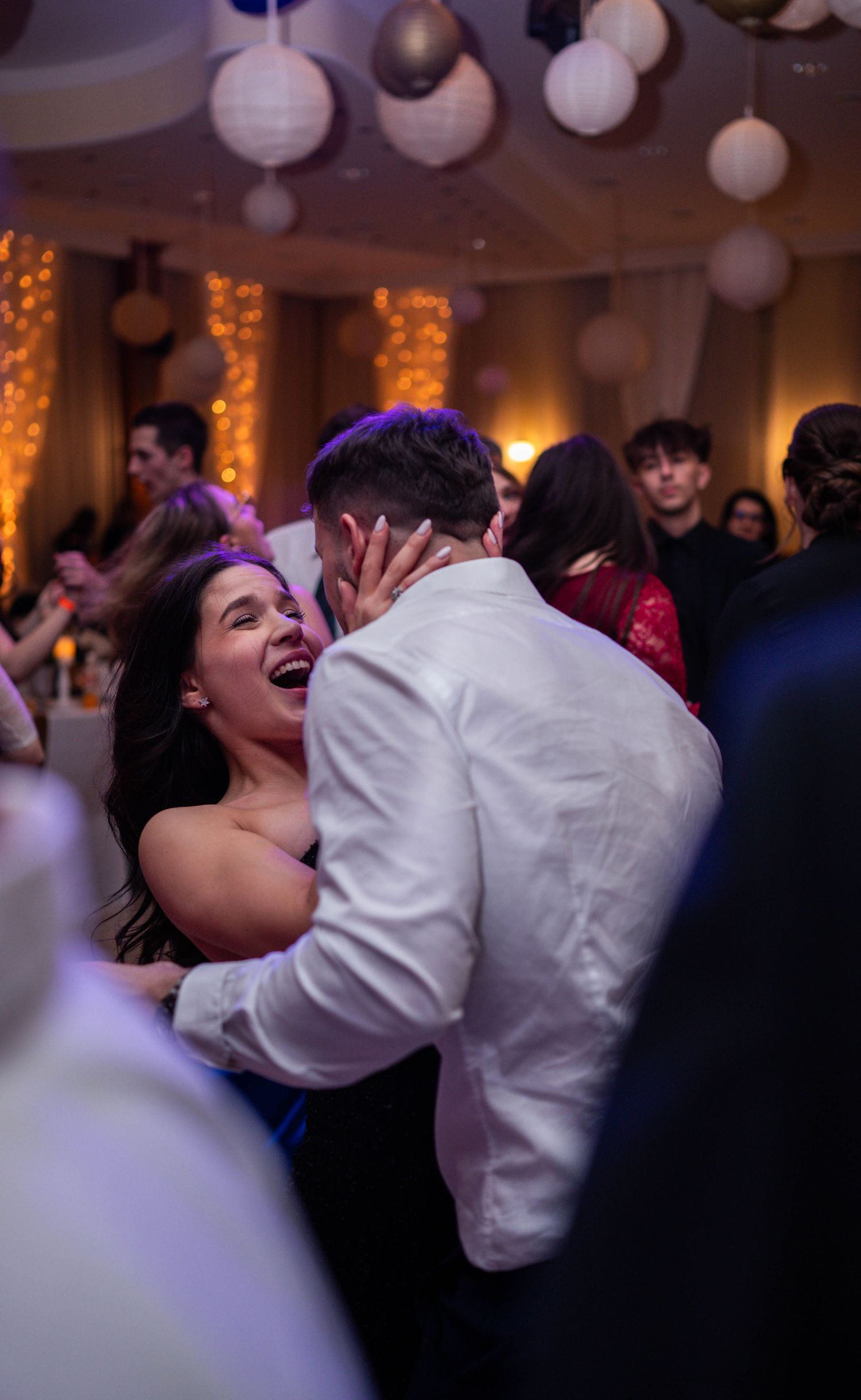Wedding guests dancing and laughing joyfully at a reception with white paper lanterns and warm lighting.