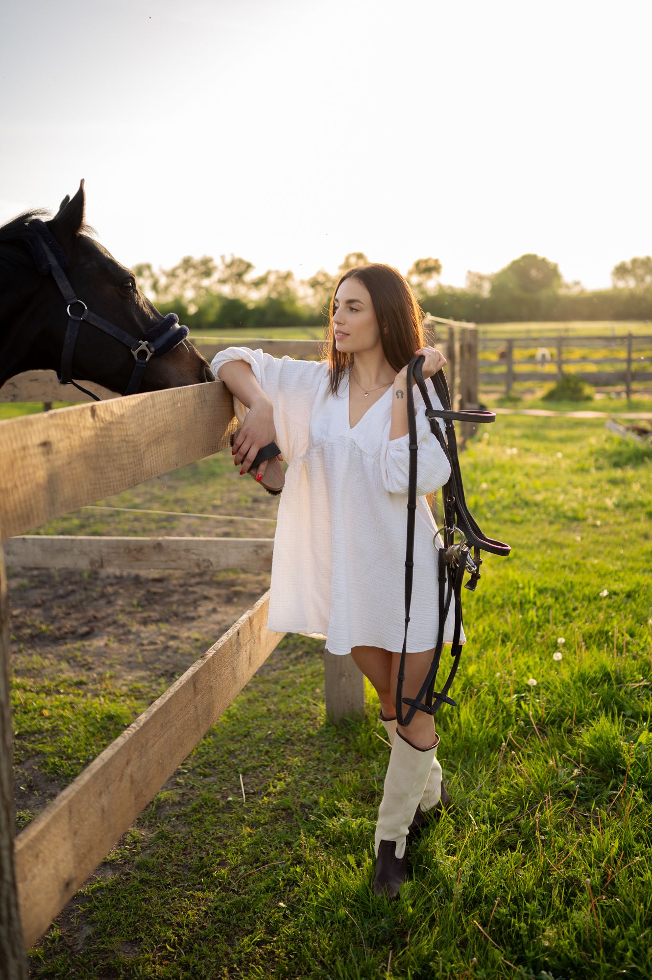 Figure in white dress leans on wooden fence near horse during sunset photoshoot.
