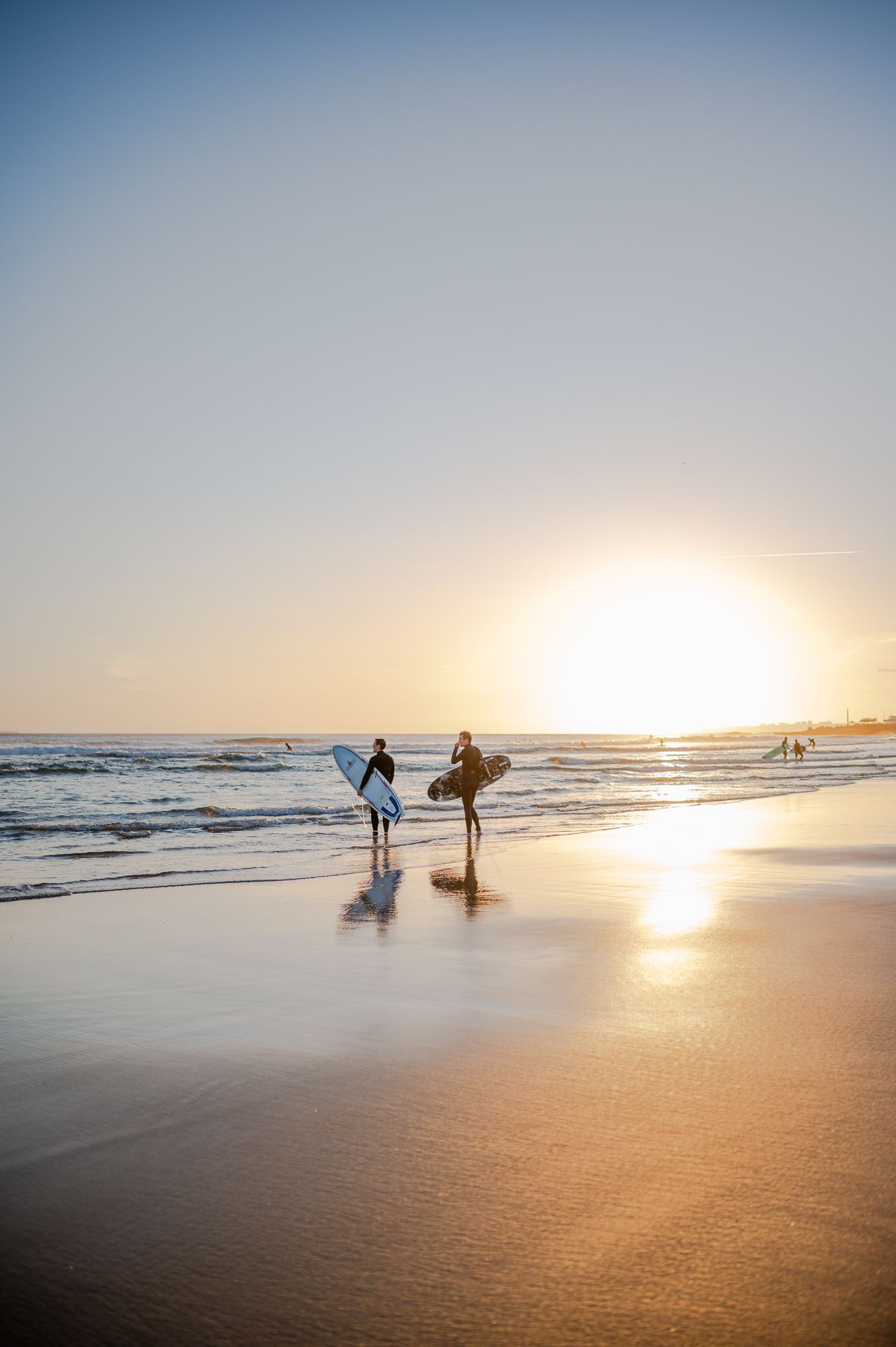Surfers walking along a golden beach at sunset with waves crashing in the distance.