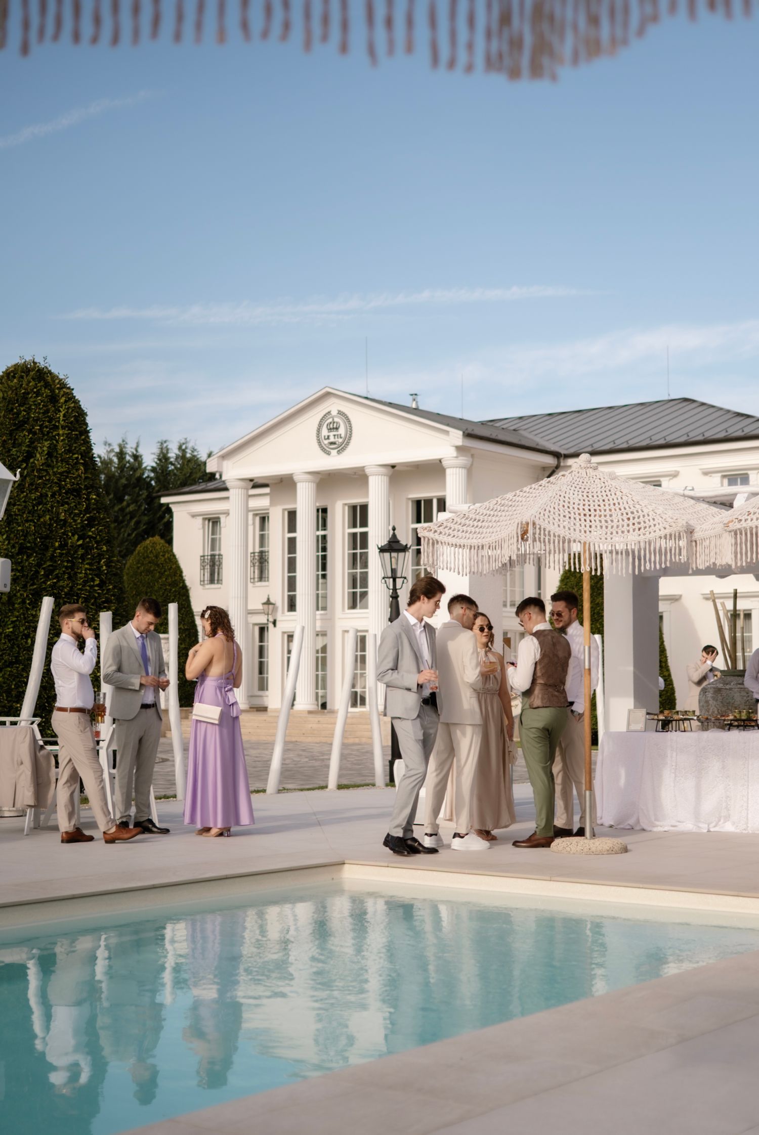 Elegant outdoor wedding reception by a pool with guests in formal attire near a white colonial-style venue.