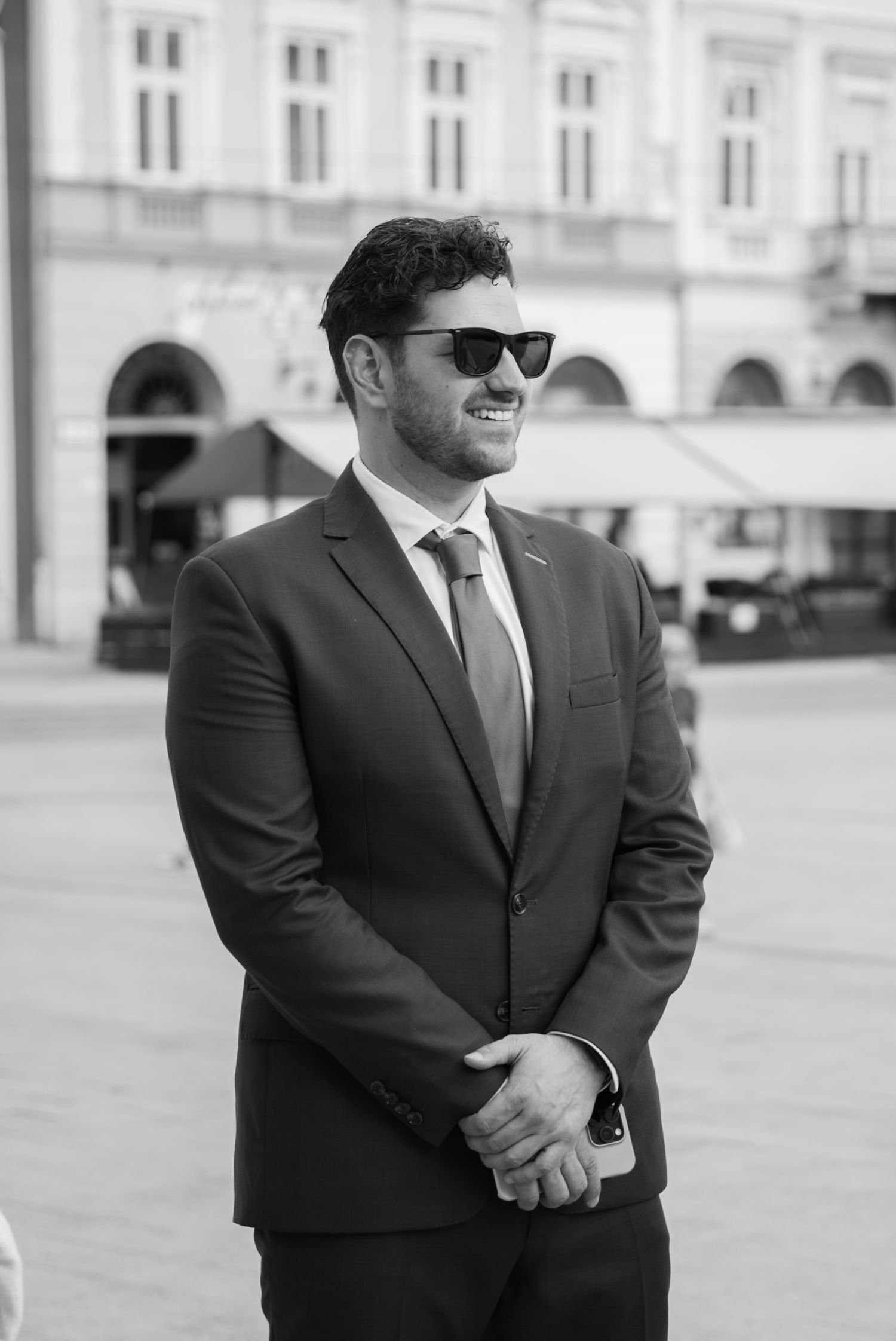 Black and white photo of a smiling man in a suit and sunglasses standing outdoors in a city square.