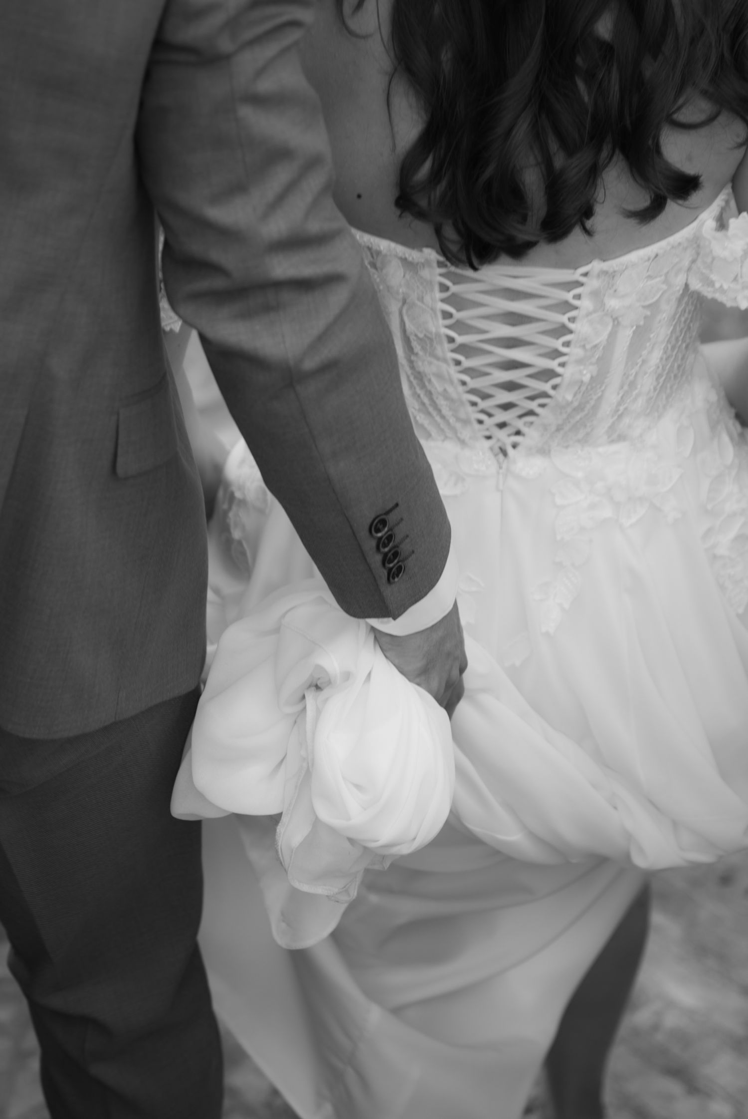 Black and white wedding photo of bride and groom holding hands, showcasing lace corset gown and groom's gray suit.