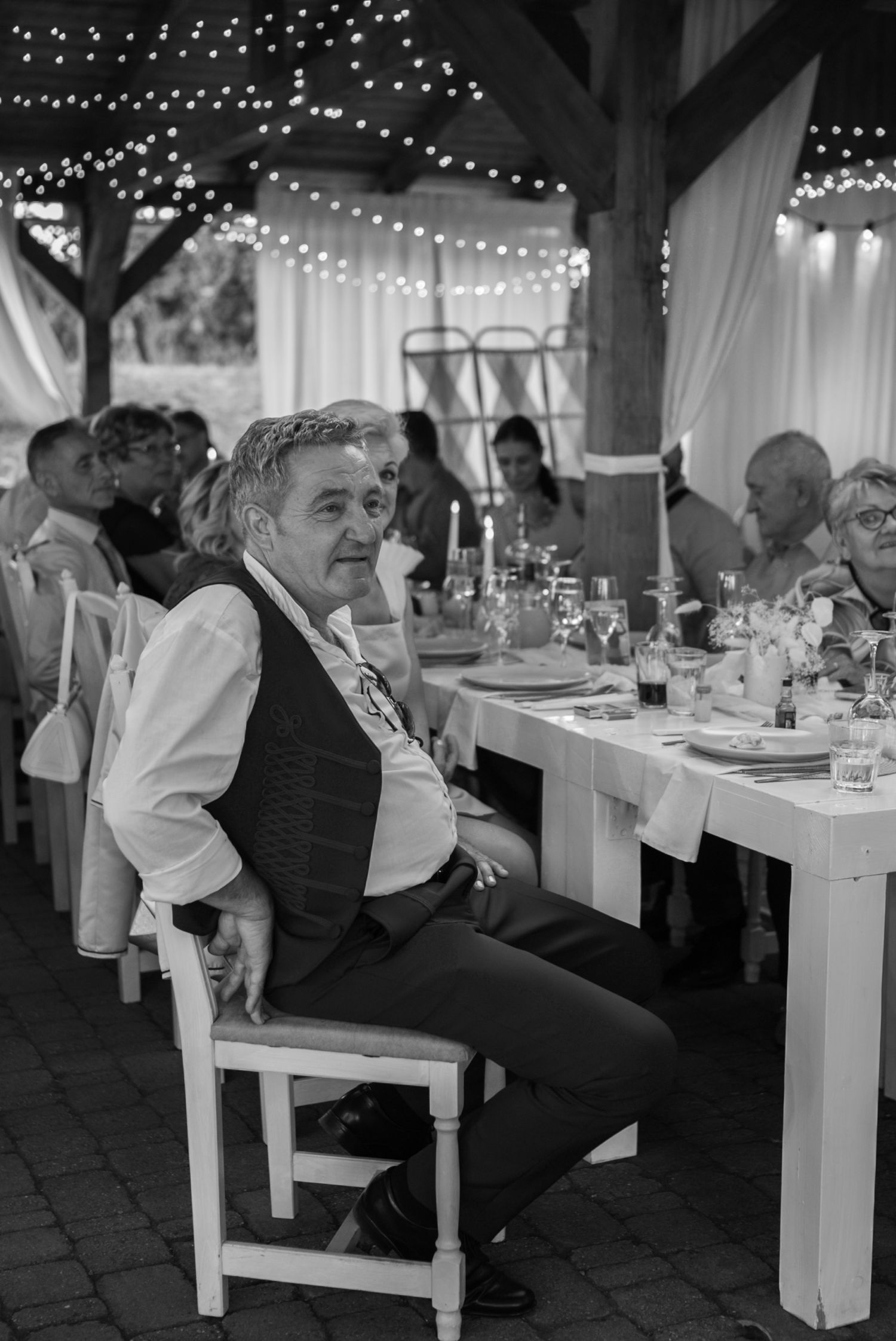 Elderly man in vest sits at decorated banquet table with string lights at formal event.