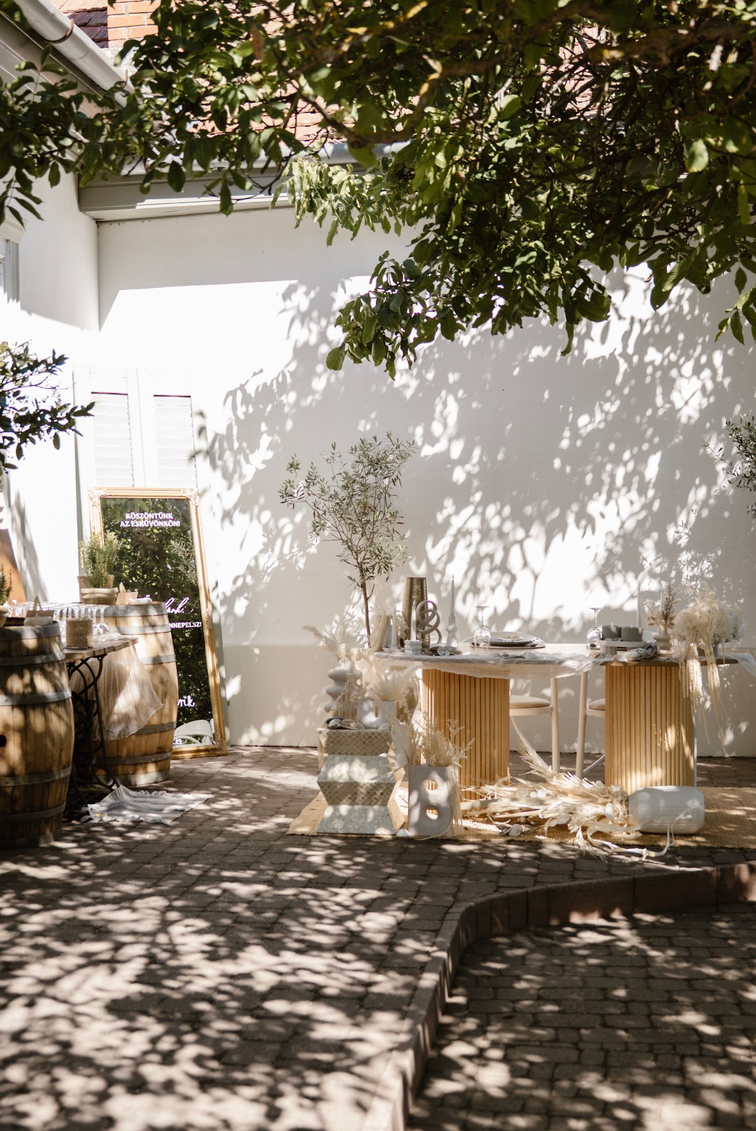 Sunlit outdoor courtyard with elegant white and gold event setup, featuring decorative trees, styled tables, and dappled shadows.