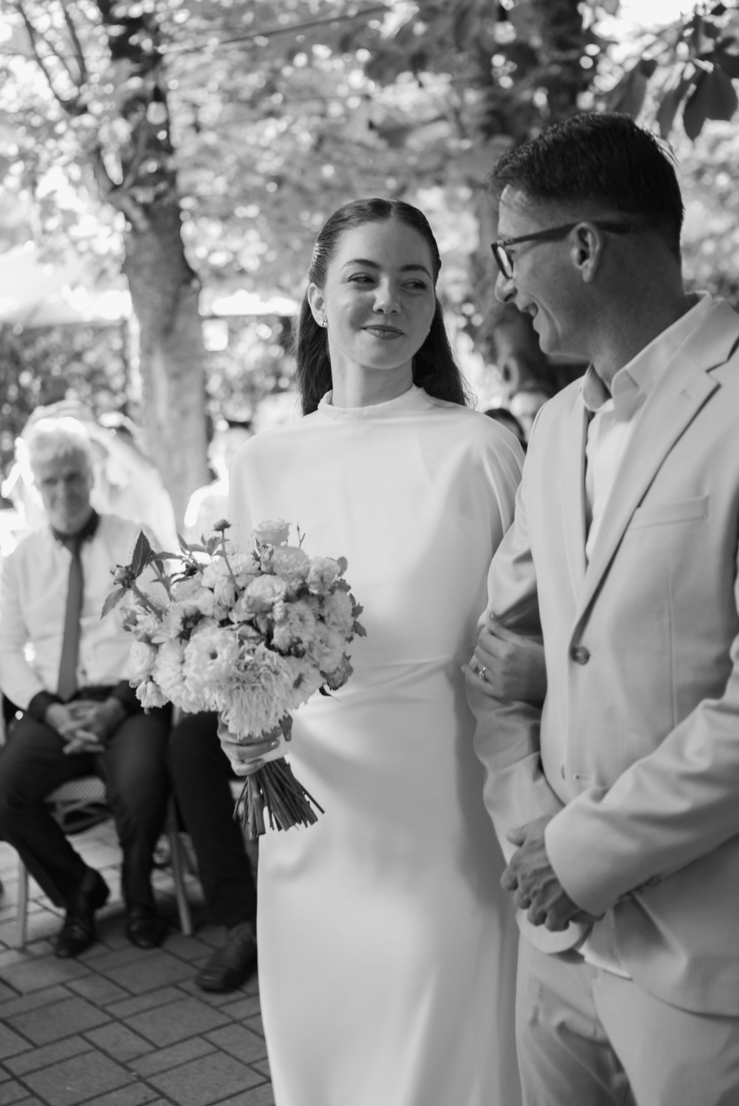 Bride in sleek white high-neck gown holding bouquet smiles at groom in light suit during outdoor wedding ceremony.