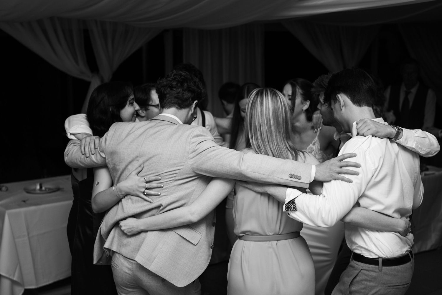 Black and white photo of wedding guests embracing in a group hug on the dance floor.