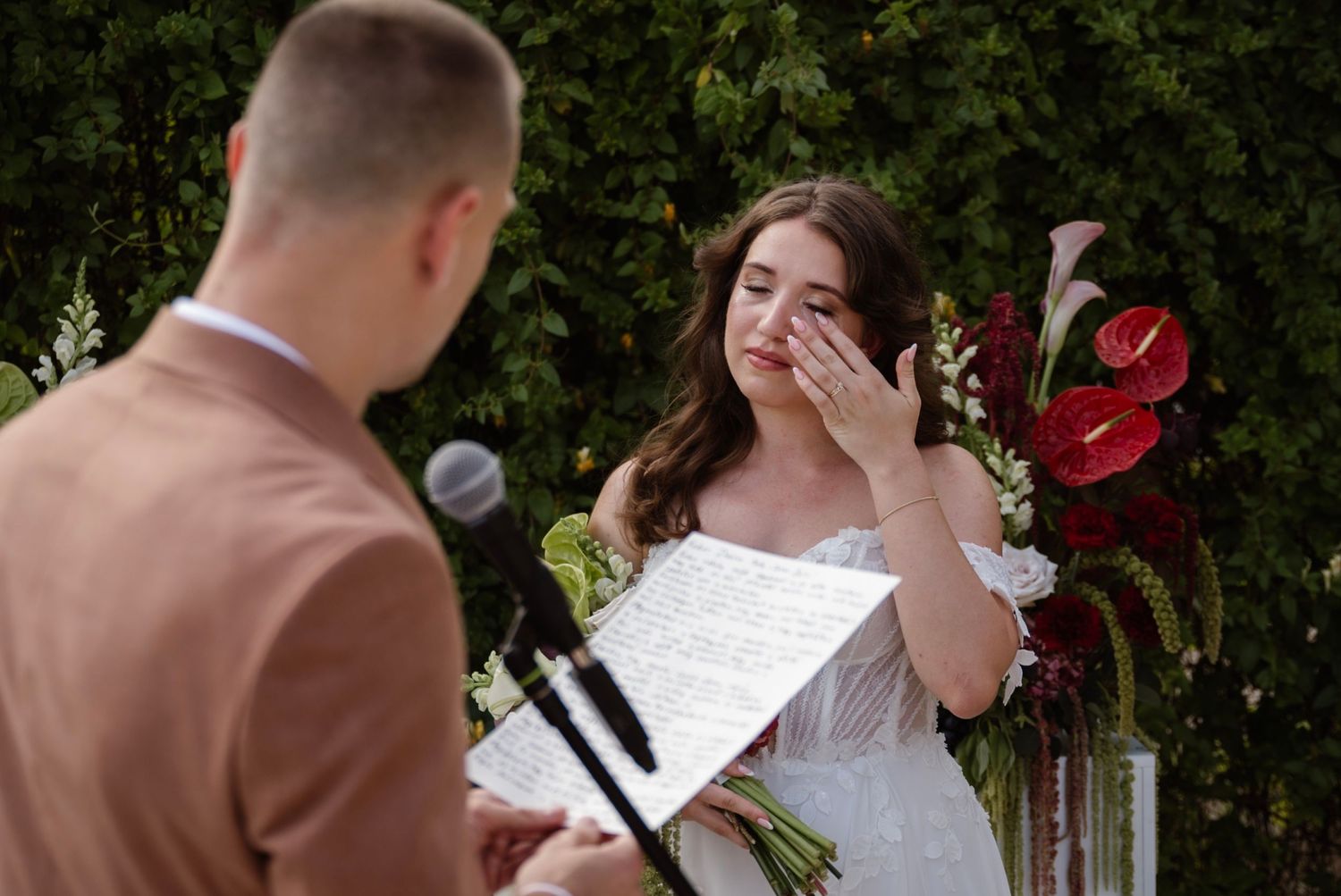Emotional bride wiping tears during outdoor wedding ceremony while holding vows, with red floral arrangement in background.