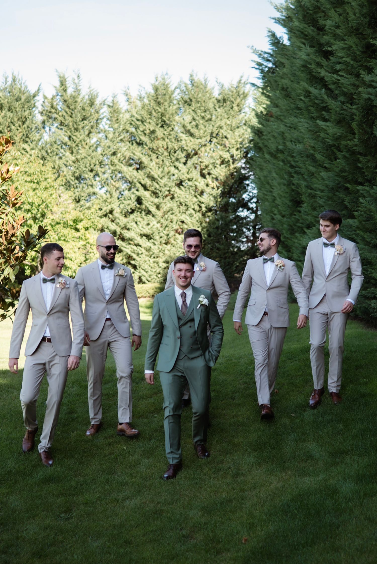 Groom in green suit walks with five groomsmen in tan suits and bow ties on a lush green lawn.