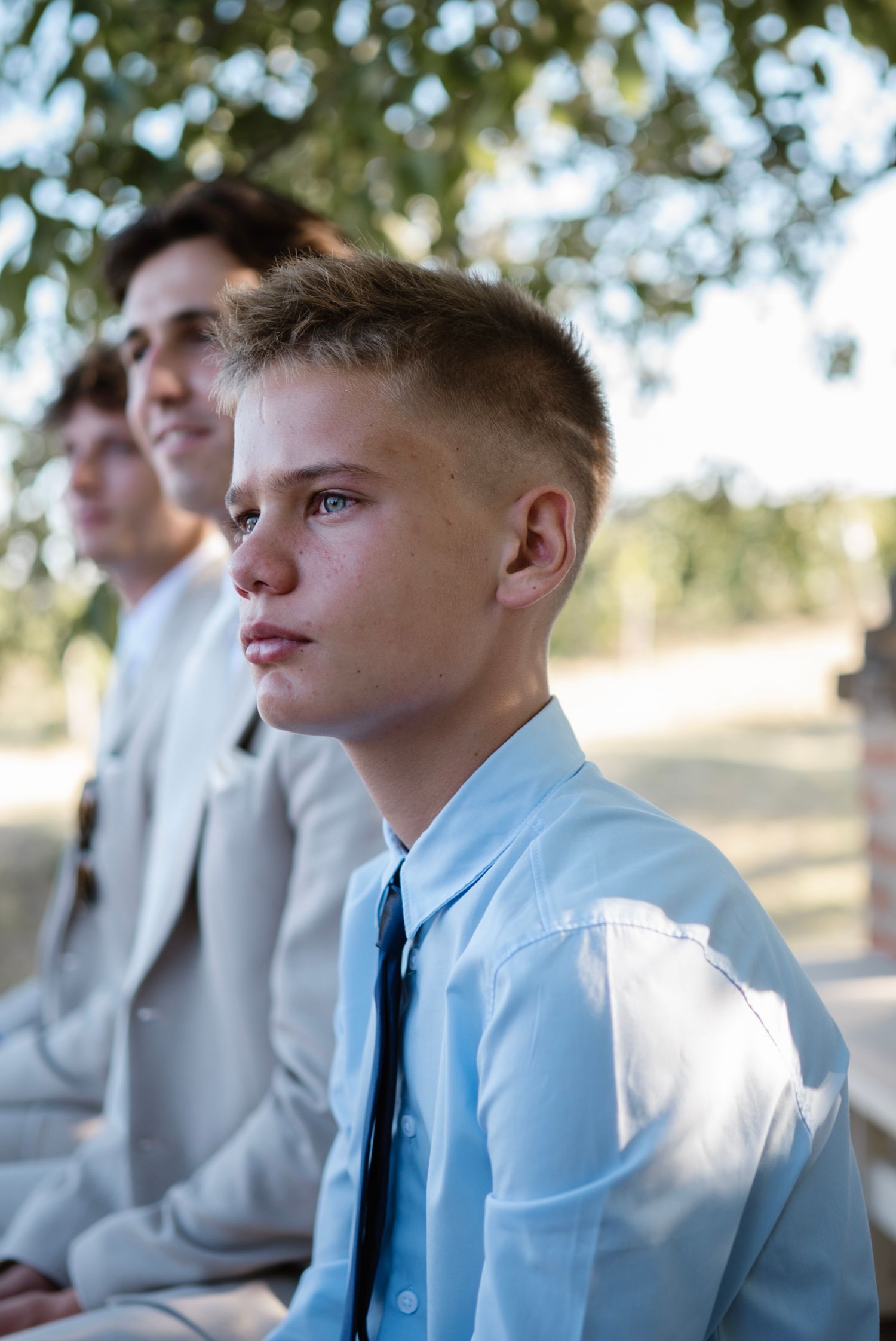 Young boy in light blue shirt and tie sitting outdoors at formal event with older teens in suits behind him.