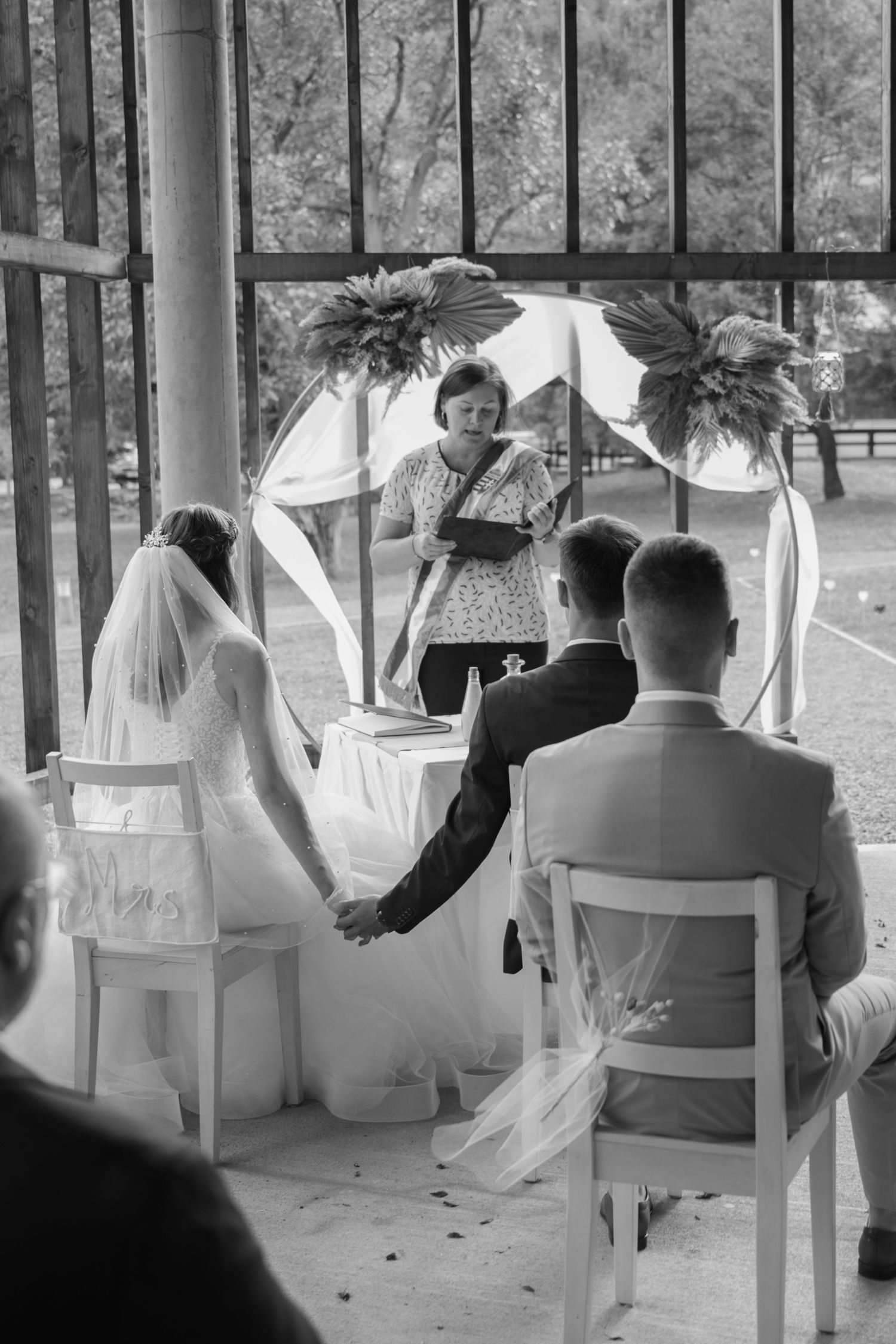 Black and white photo of a wedding ceremony under a decorated arch with white chairs.
