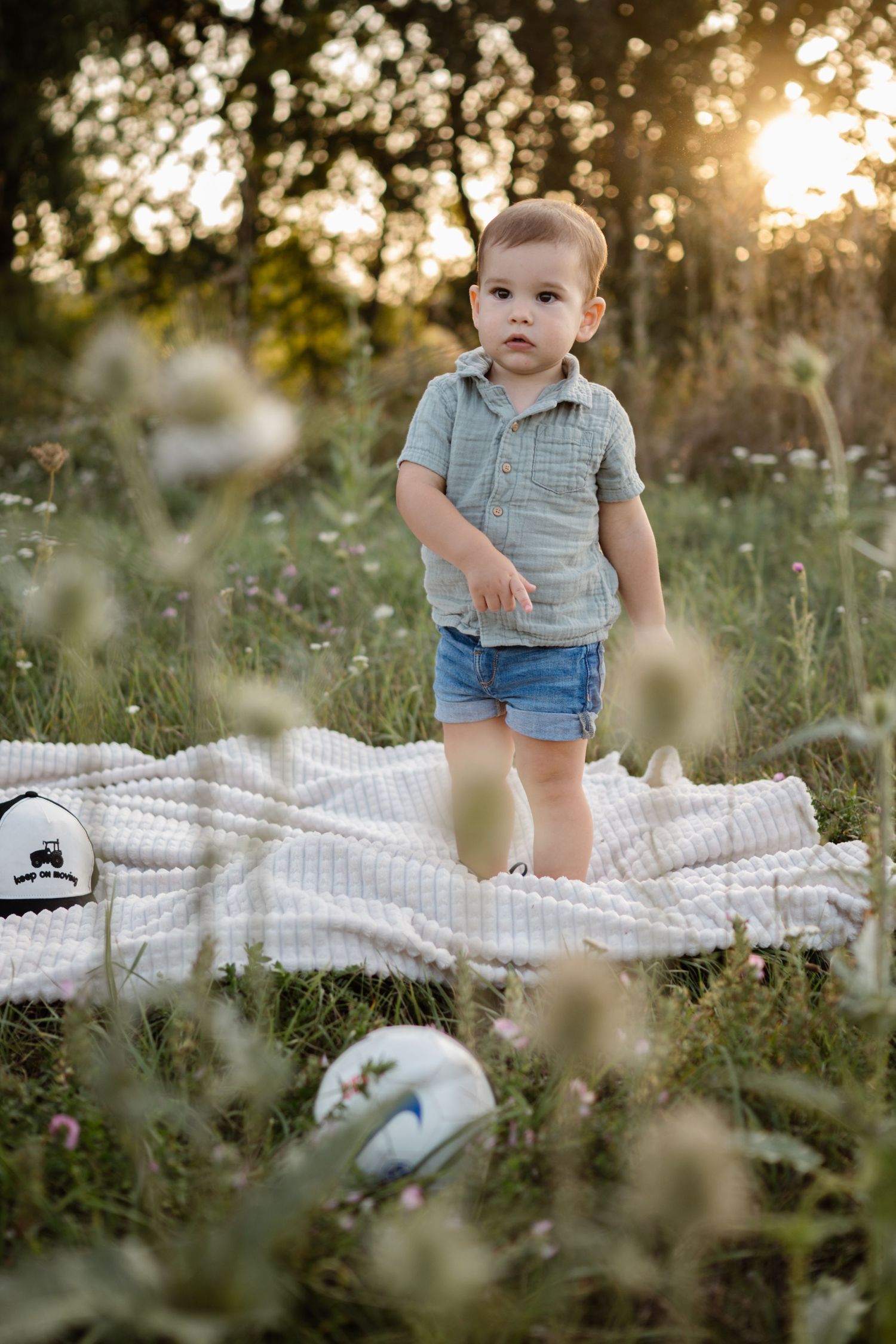 A toddler stands on a picnic blanket in a field at sunset wearing a grey polo shirt and blue shorts.