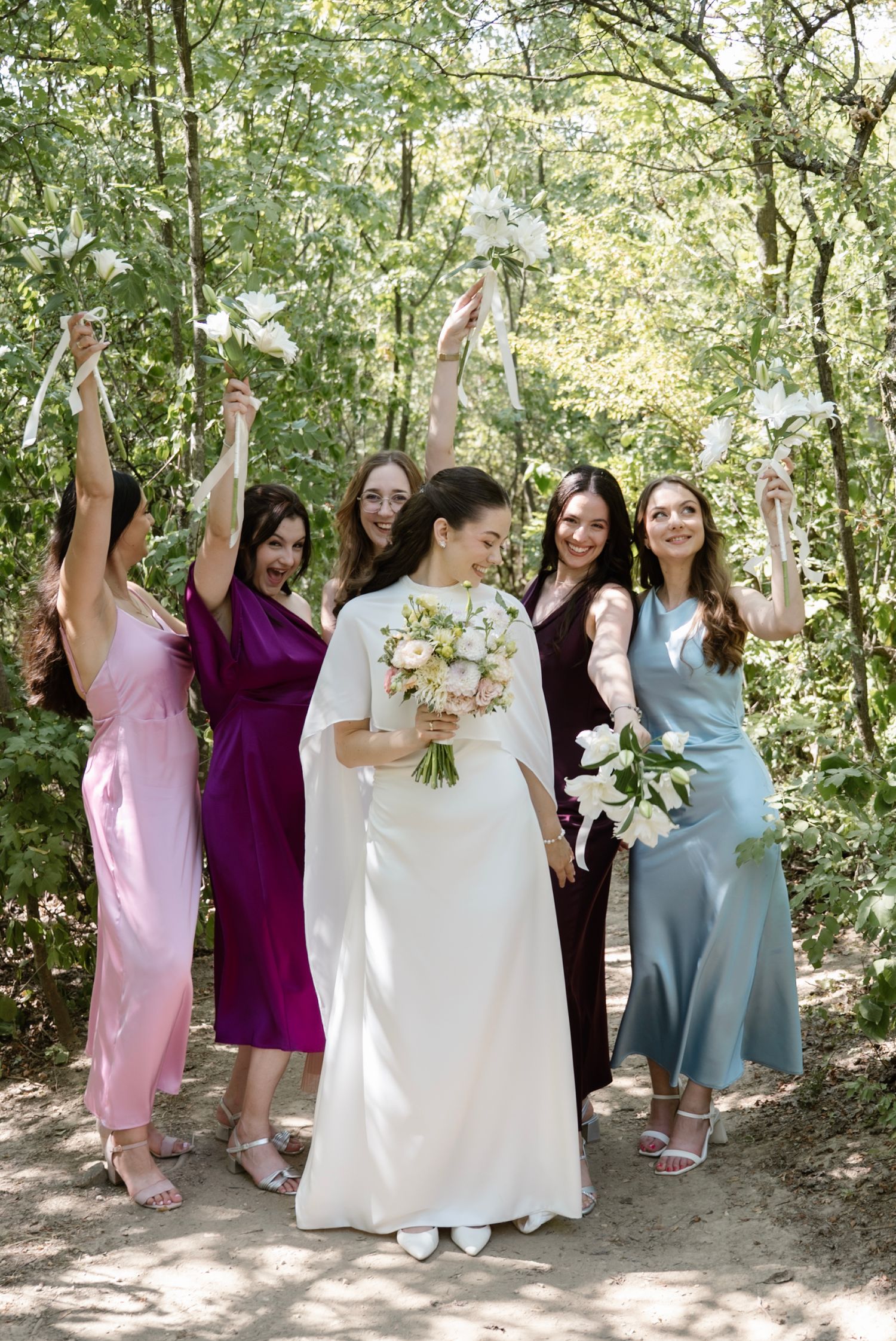 Bride in white gown holds floral bouquet with bridesmaids in colorful dresses celebrating outdoors in a forest setting.
