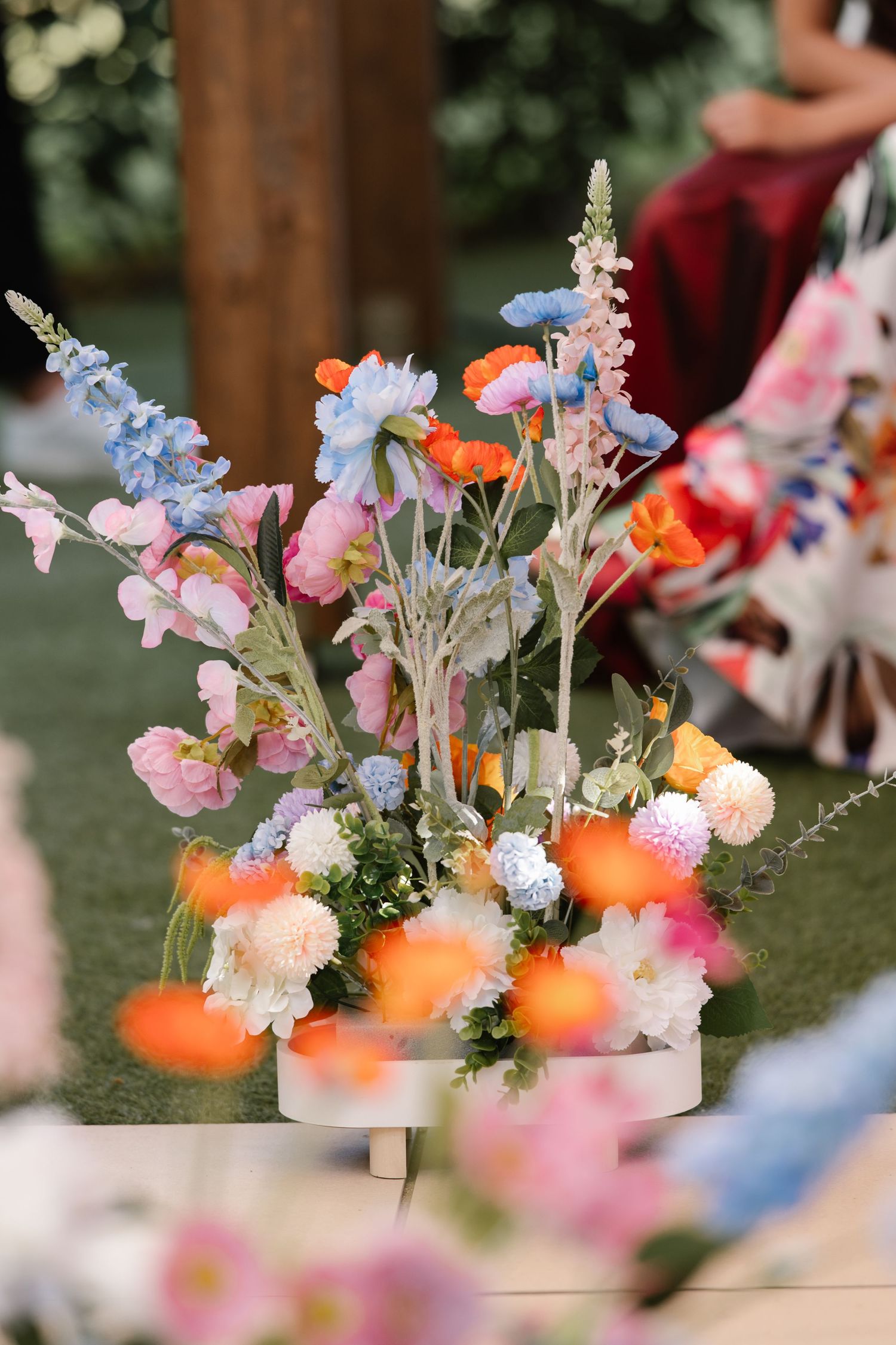 Delicate floral arrangements with pink, orange, and blue flowers displayed on white pedestals in an outdoor garden setting.
