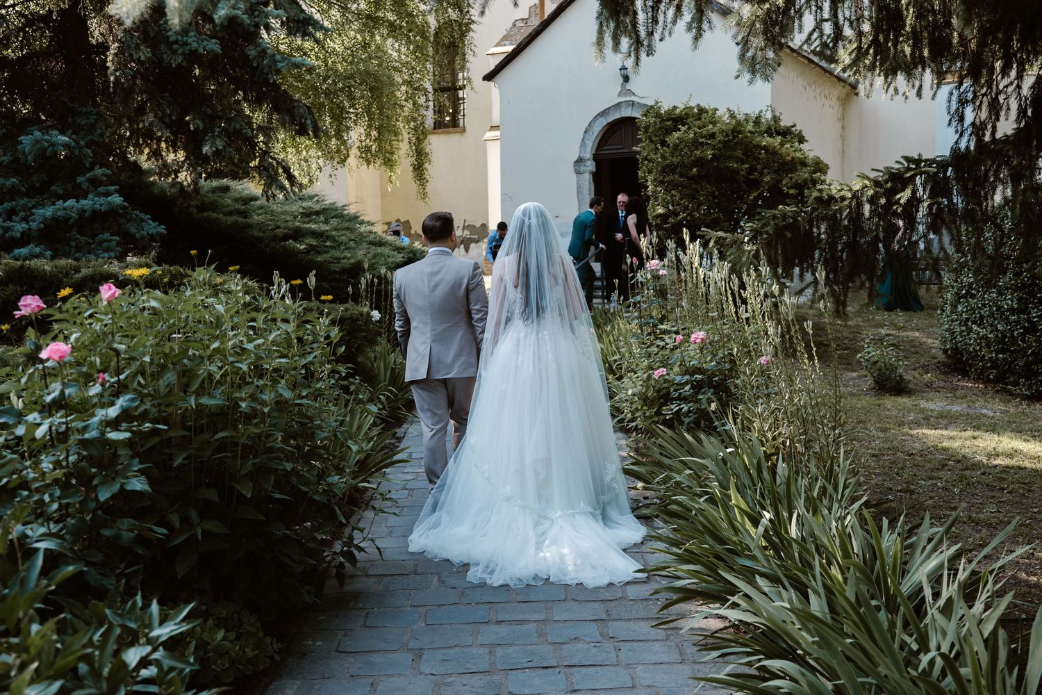 A couple walks down a garden path towards a church entrance.