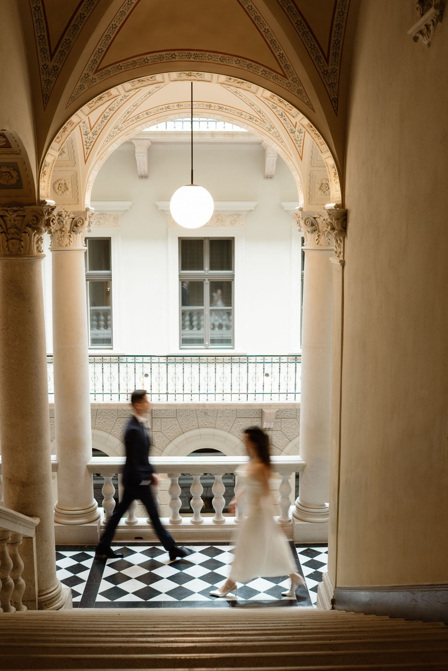 Bride and groom walk through elegant arched hallway with checkered floors and ornate columns in a grand historic building.