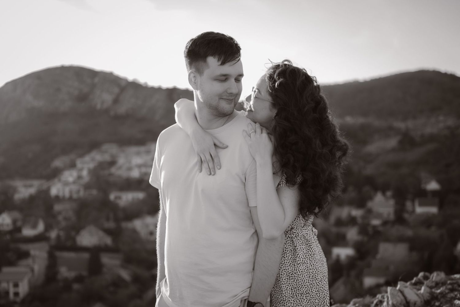 Black and white photo of a romantic couple sharing a tender moment with scenic hills and village in the background.