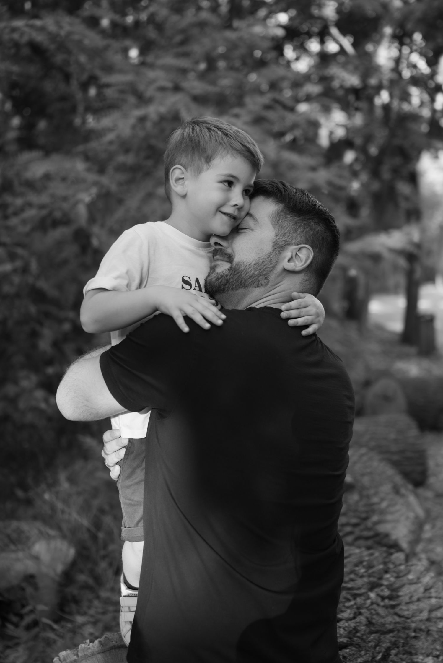 Black and white photo of a bearded man lovingly holding a young smiling boy outdoors among trees.