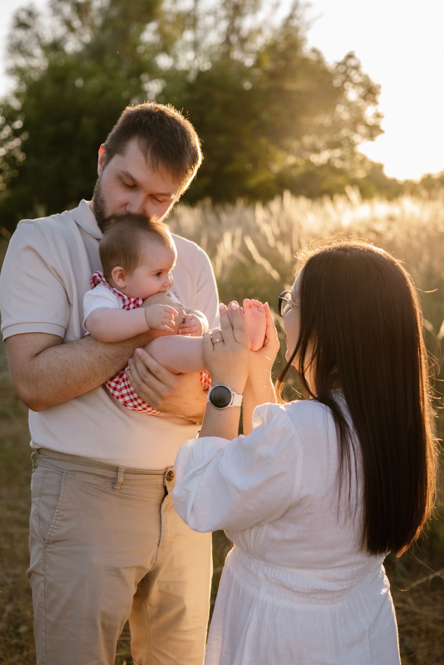 A young family enjoys a golden sunset outdoors, father holding baby while mother kisses the infant's tiny feet.