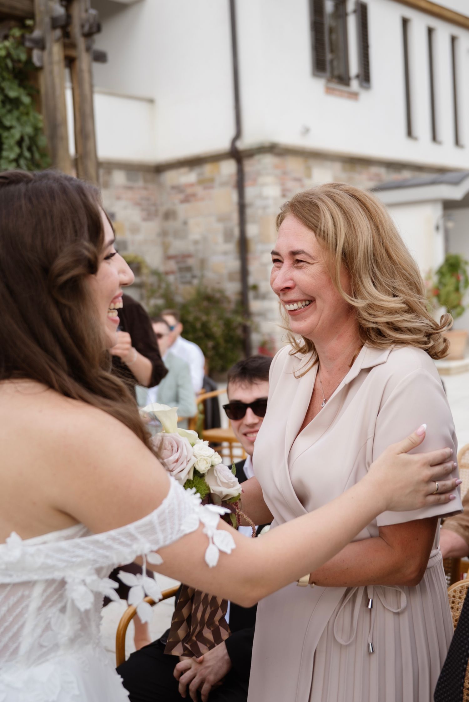 Bride in off-shoulder lace gown laughing and embracing a smiling blonde woman guest at outdoor wedding.