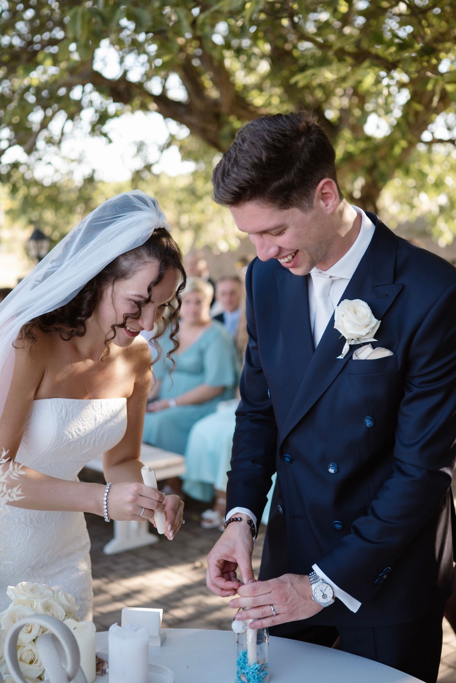 Bride and groom smiling during sand unity ceremony at outdoor wedding reception under trees.