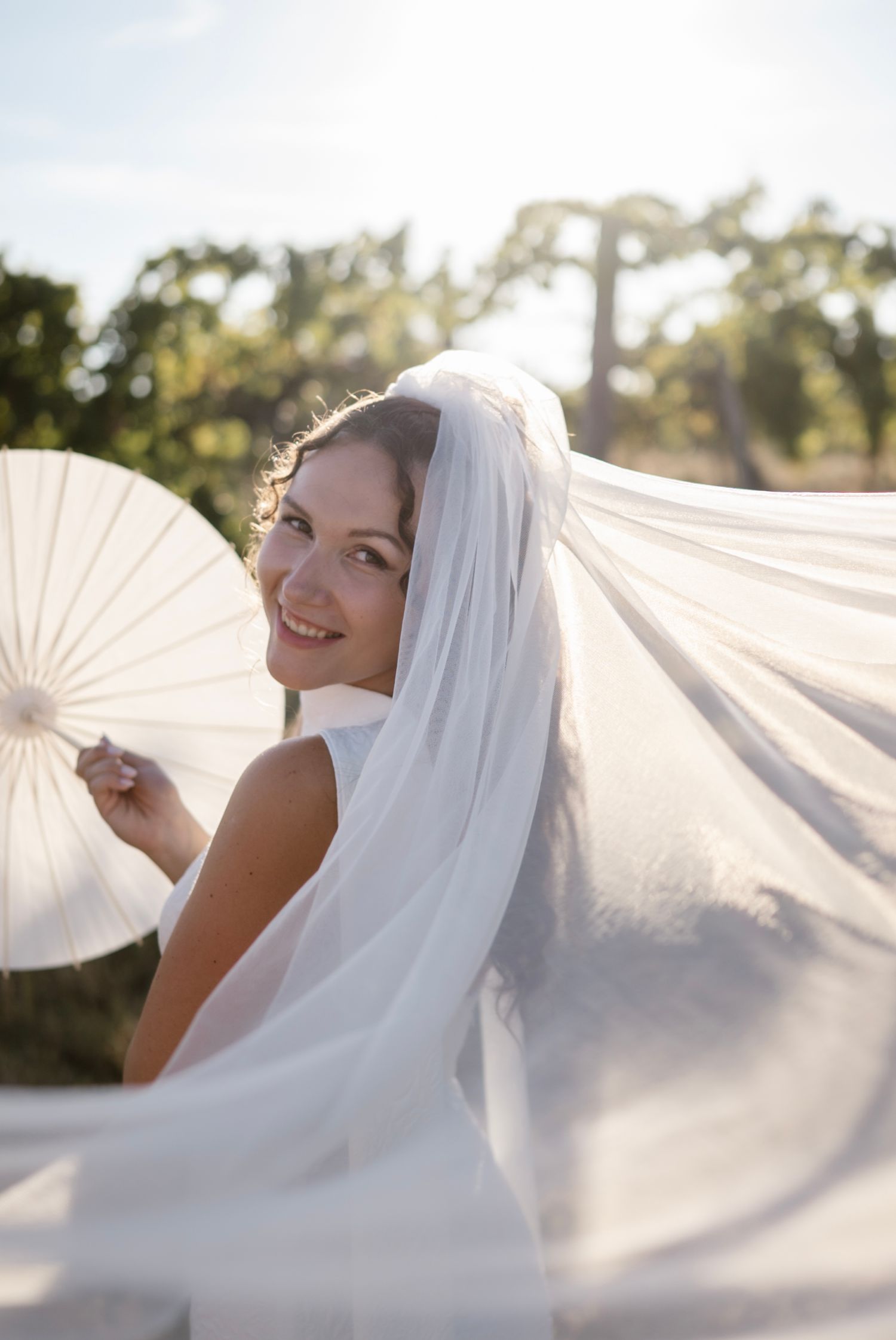 Smiling bride with flowing veil holding a white parasol outdoors in sunlight.