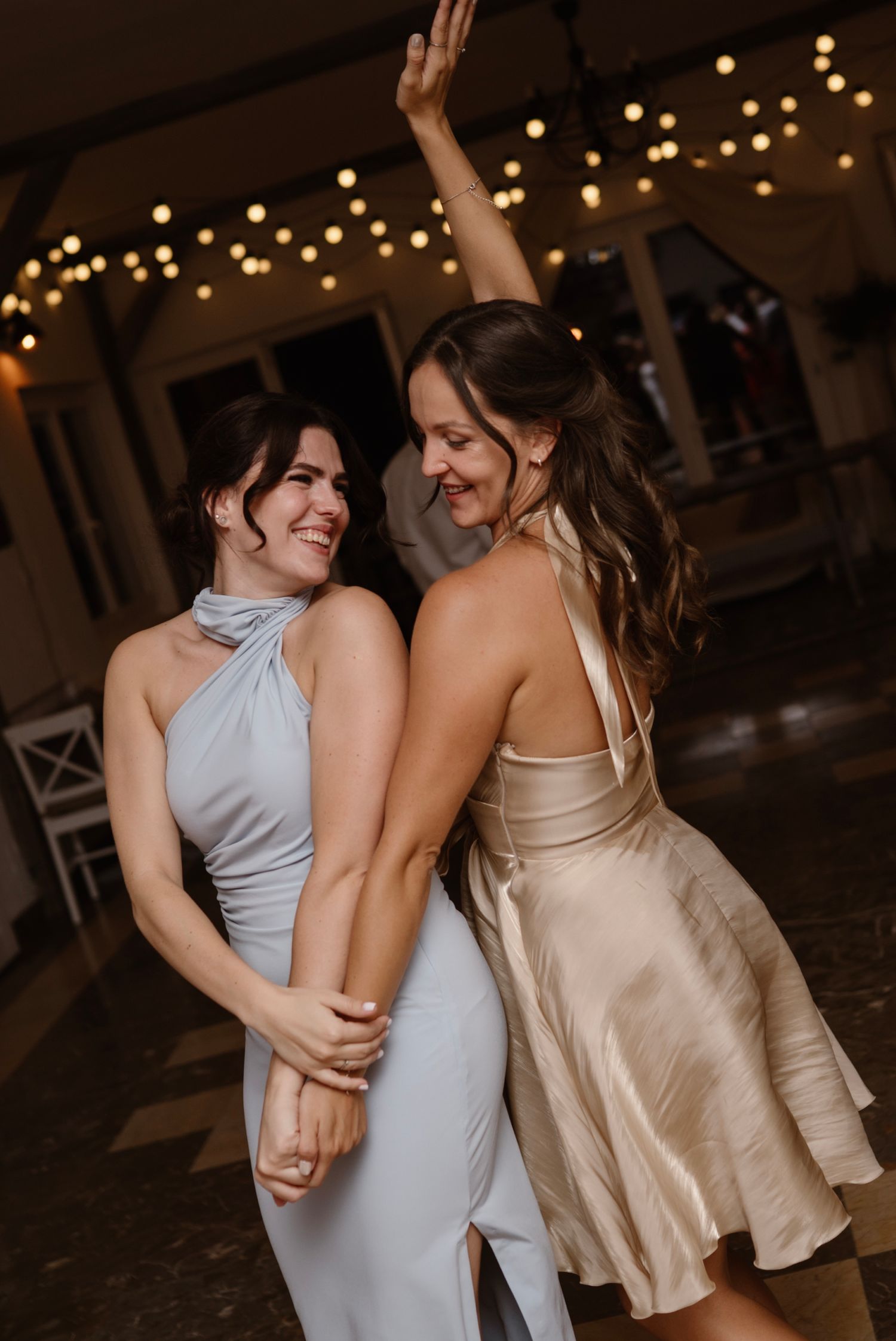 Two women in elegant dresses dancing joyfully at wedding reception with string lights backdrop.