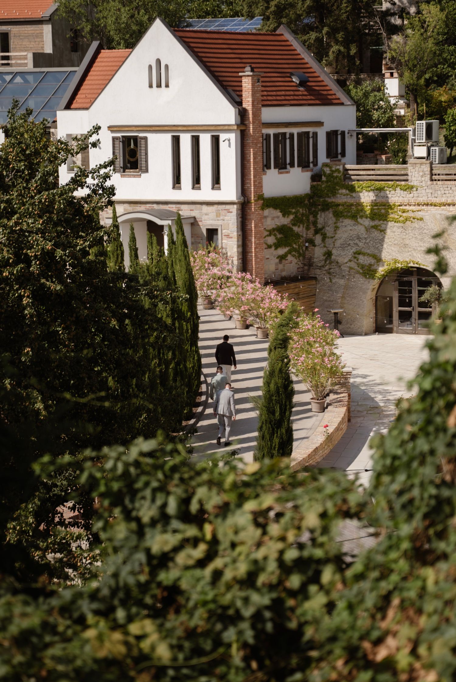 Two people walk along a stone pathway beside a charming white villa with terracotta roof surrounded by lush greenery.