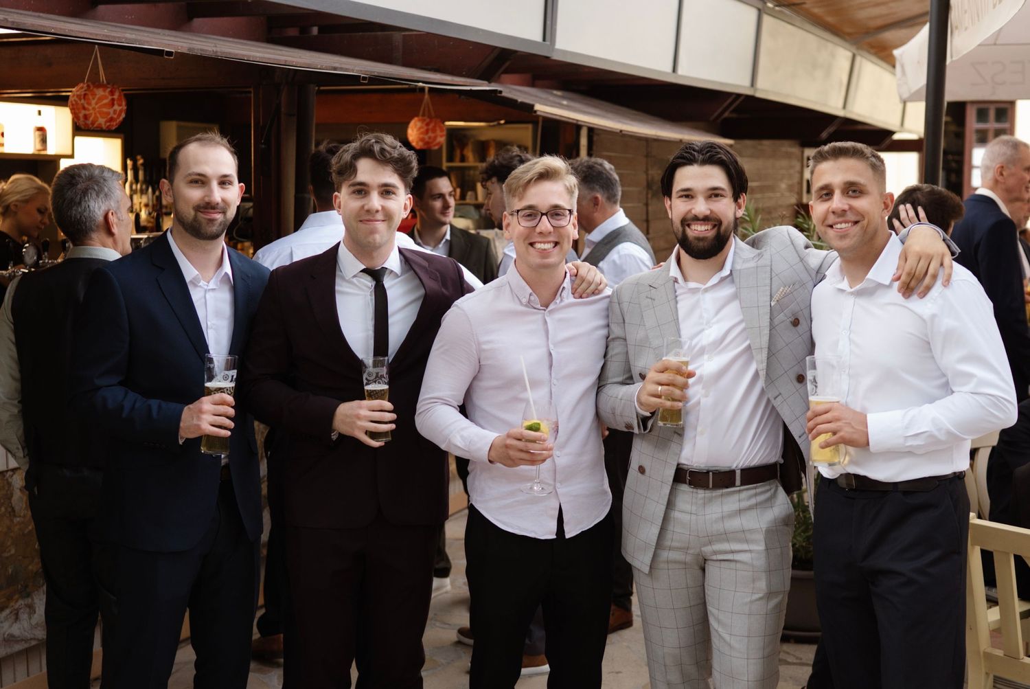Five smiling men in formal attire pose together holding drinks at an outdoor wedding reception.