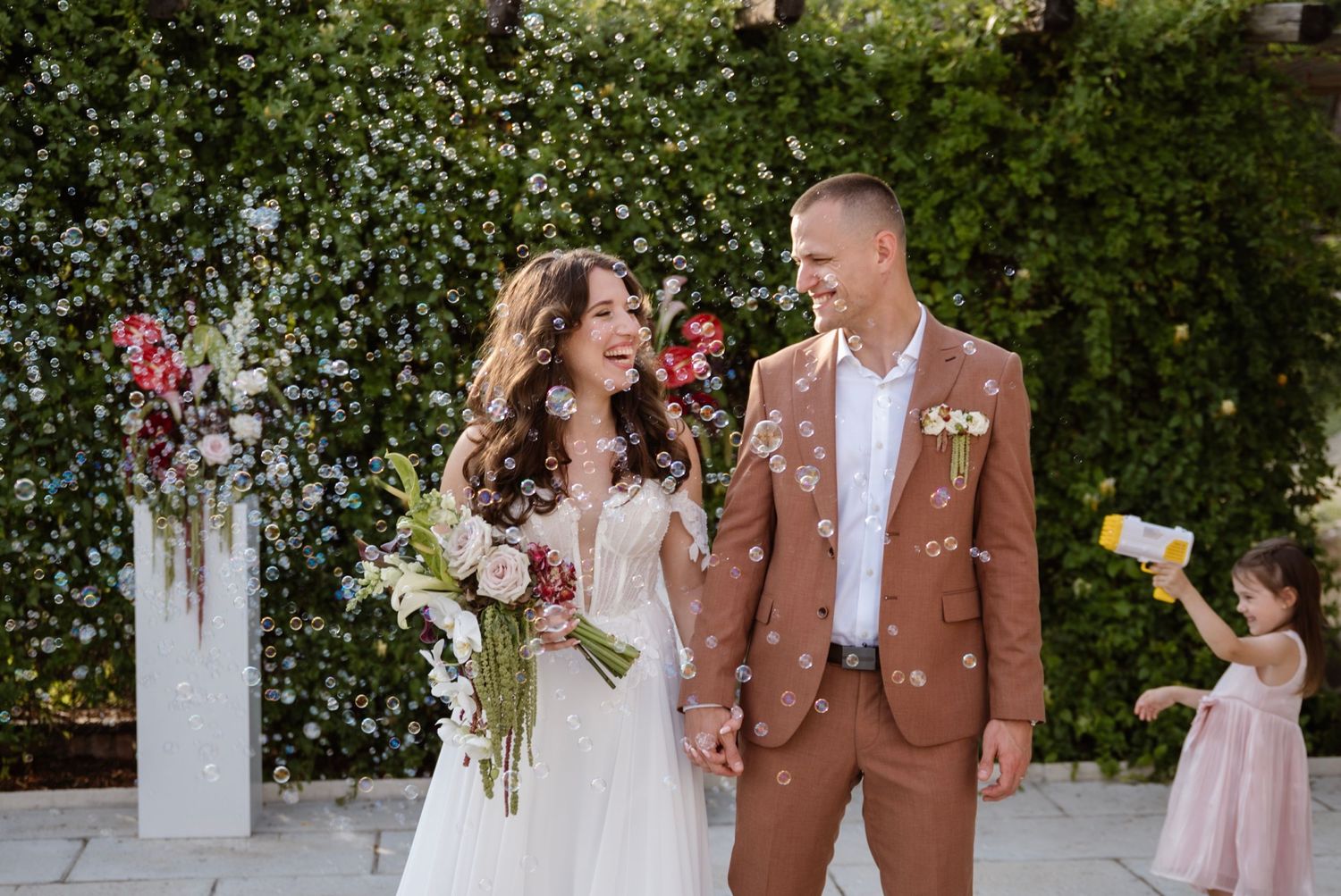Happy newlyweds laughing amid bubbles outdoors, bride in white gown holding bouquet, groom in brown suit, flower girl nearby.